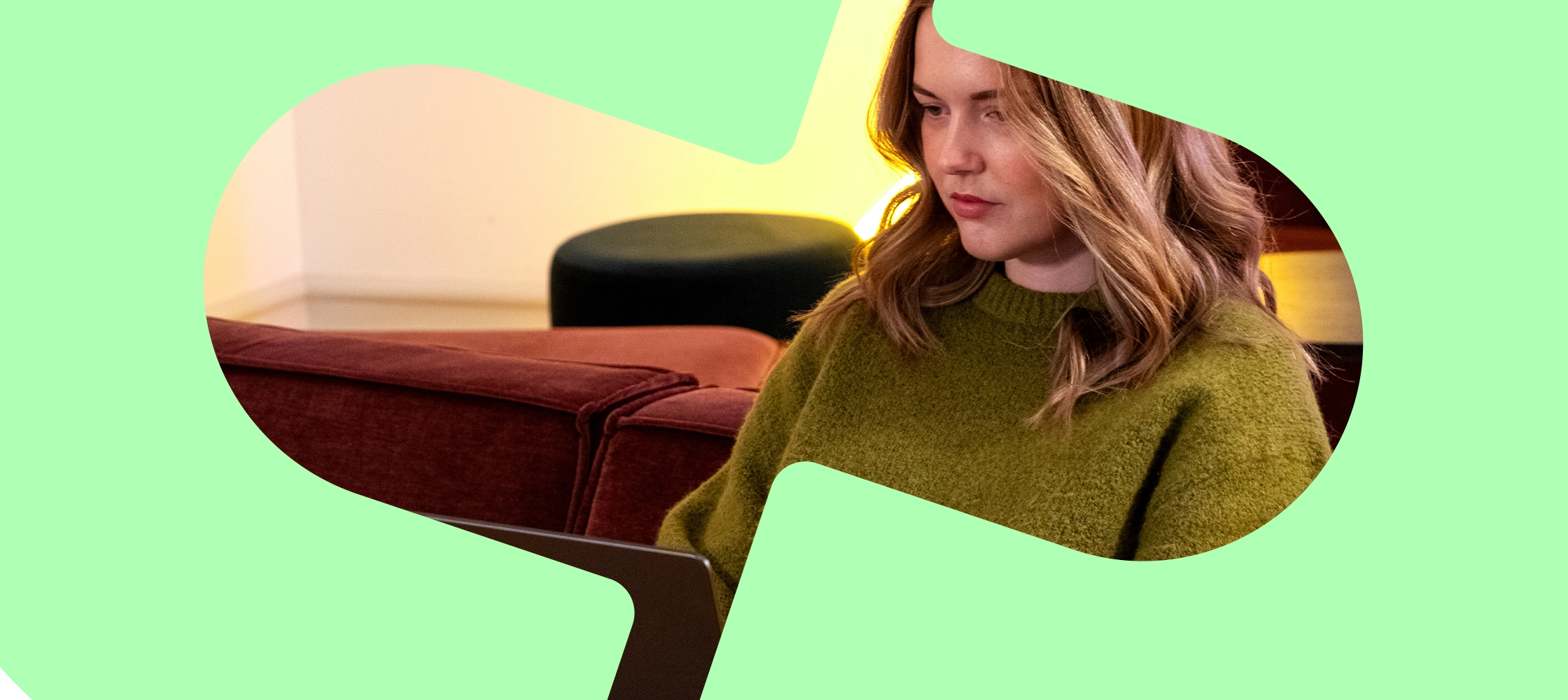 Kvinna i en grön tröja som arbetar på en bärbar dator på en vinröd soffa mot en grön bakgrund.