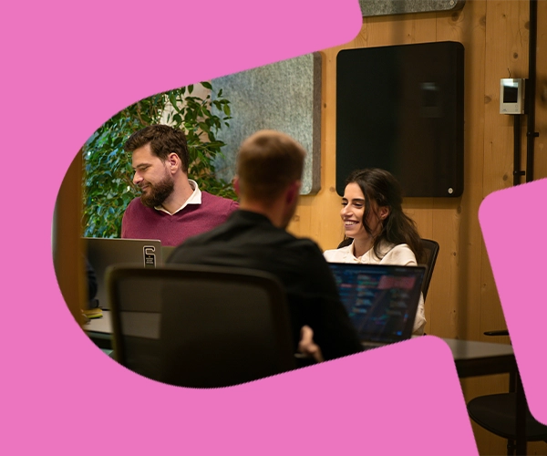 Tre kollegor diskuterar Employer Marketing-strategi vid laptops på ett modernt kontor, inramade av en rosa grafisk form.