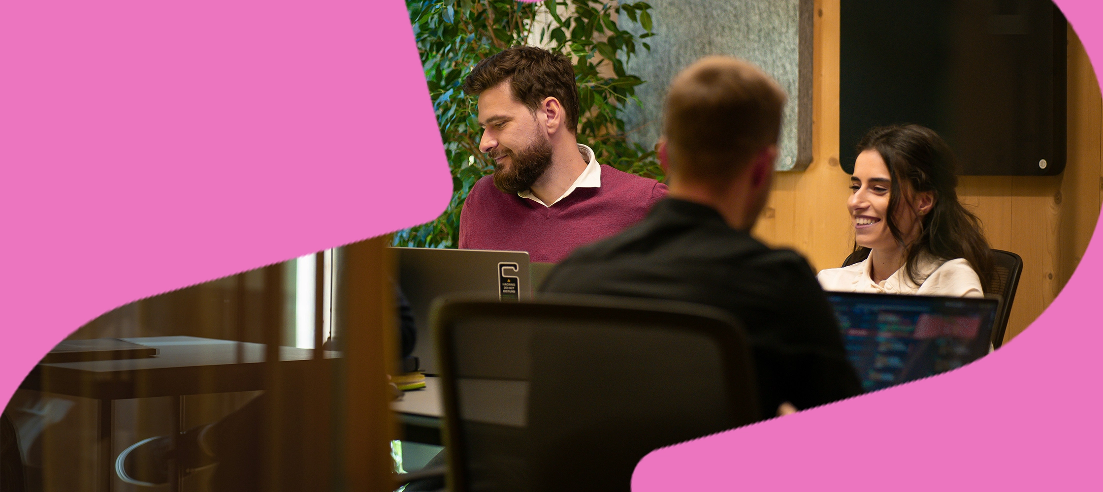Three colleagues discussing Employer Marketing strategy around laptops in a modern office, framed by a pink graphic shape.