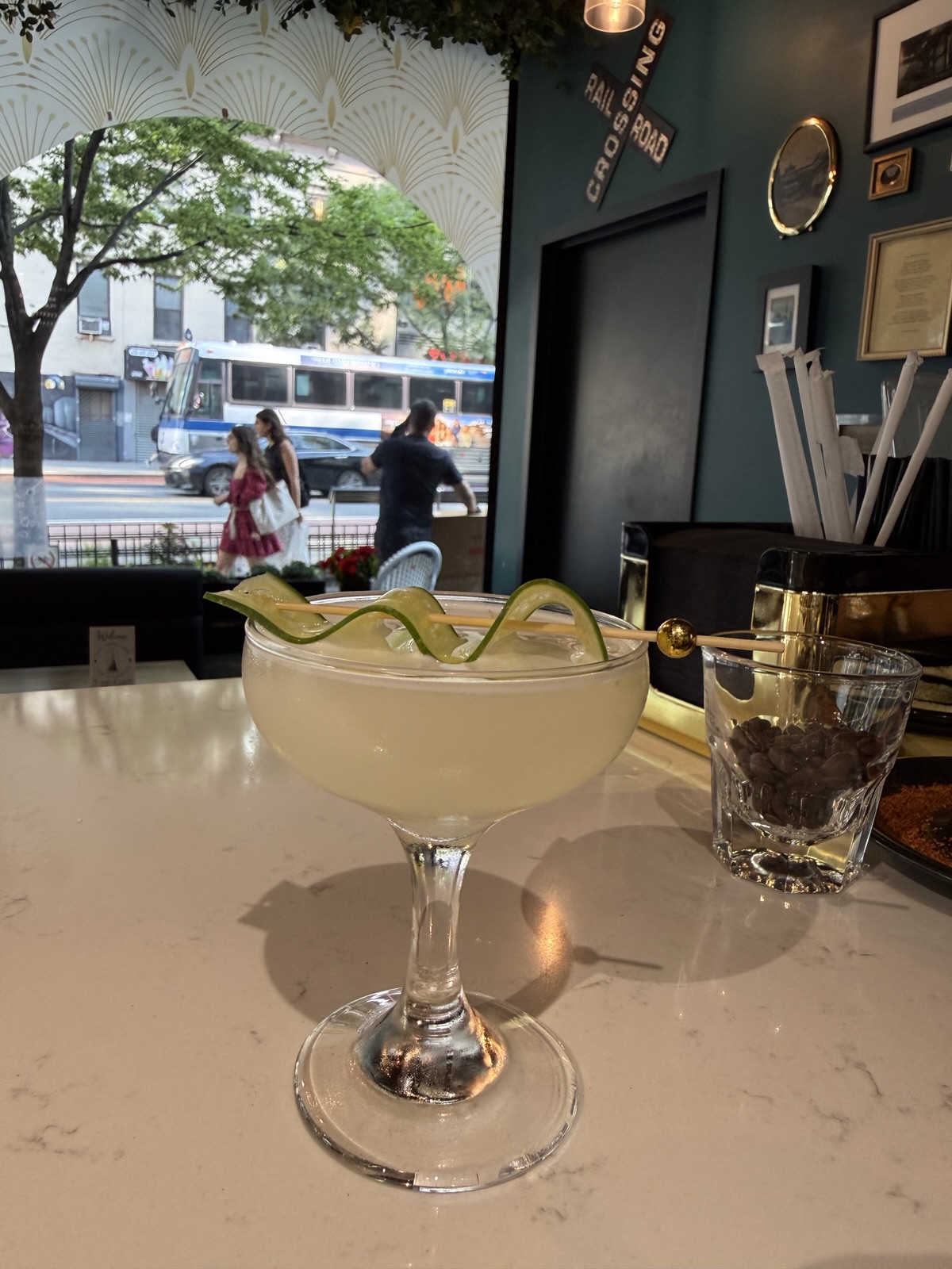 Murray Hill Bloom cocktail in a coupe with a cucumber ribbon on a gold pick, beneath a vintage railroad crossing sign, an MTA bus passing on 2nd Avenue outside