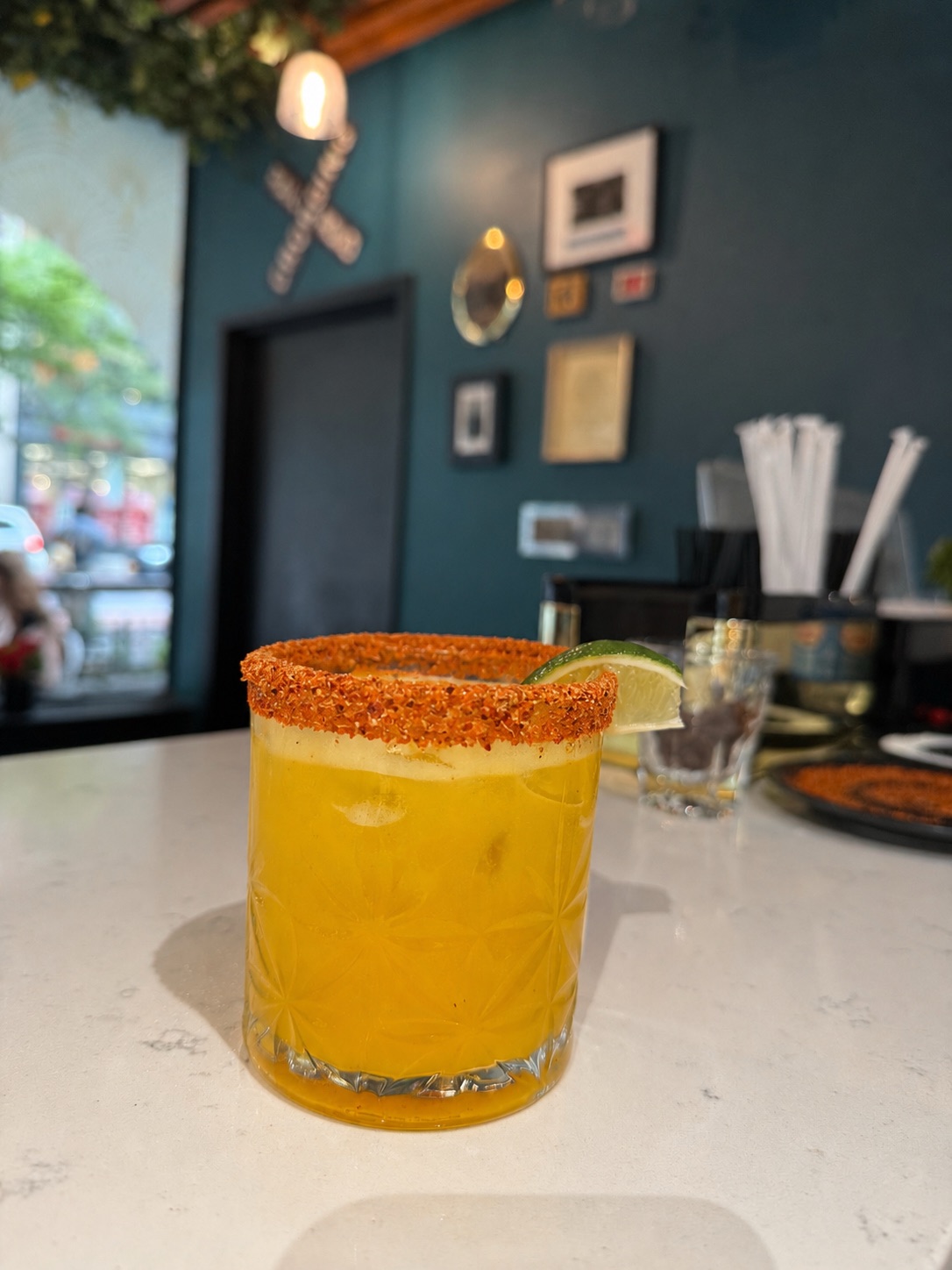 Spicy Mango Fizz mocktail with a chili-salt rim and lime wedge on the bar, against the cafe teal wall and gallery frames