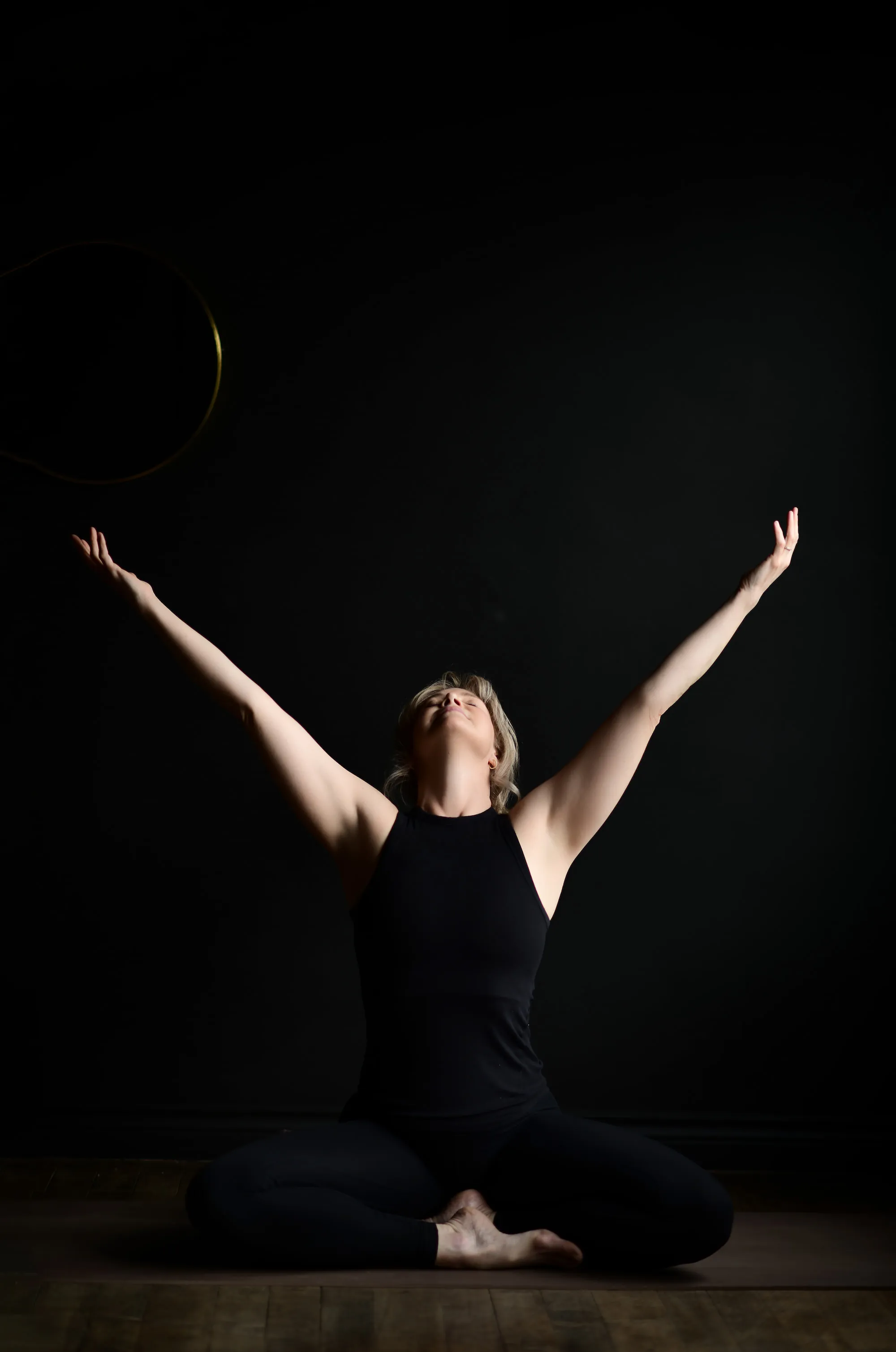 Woman dressed in black sitting crossed legged, with arms outstretched to sky. 