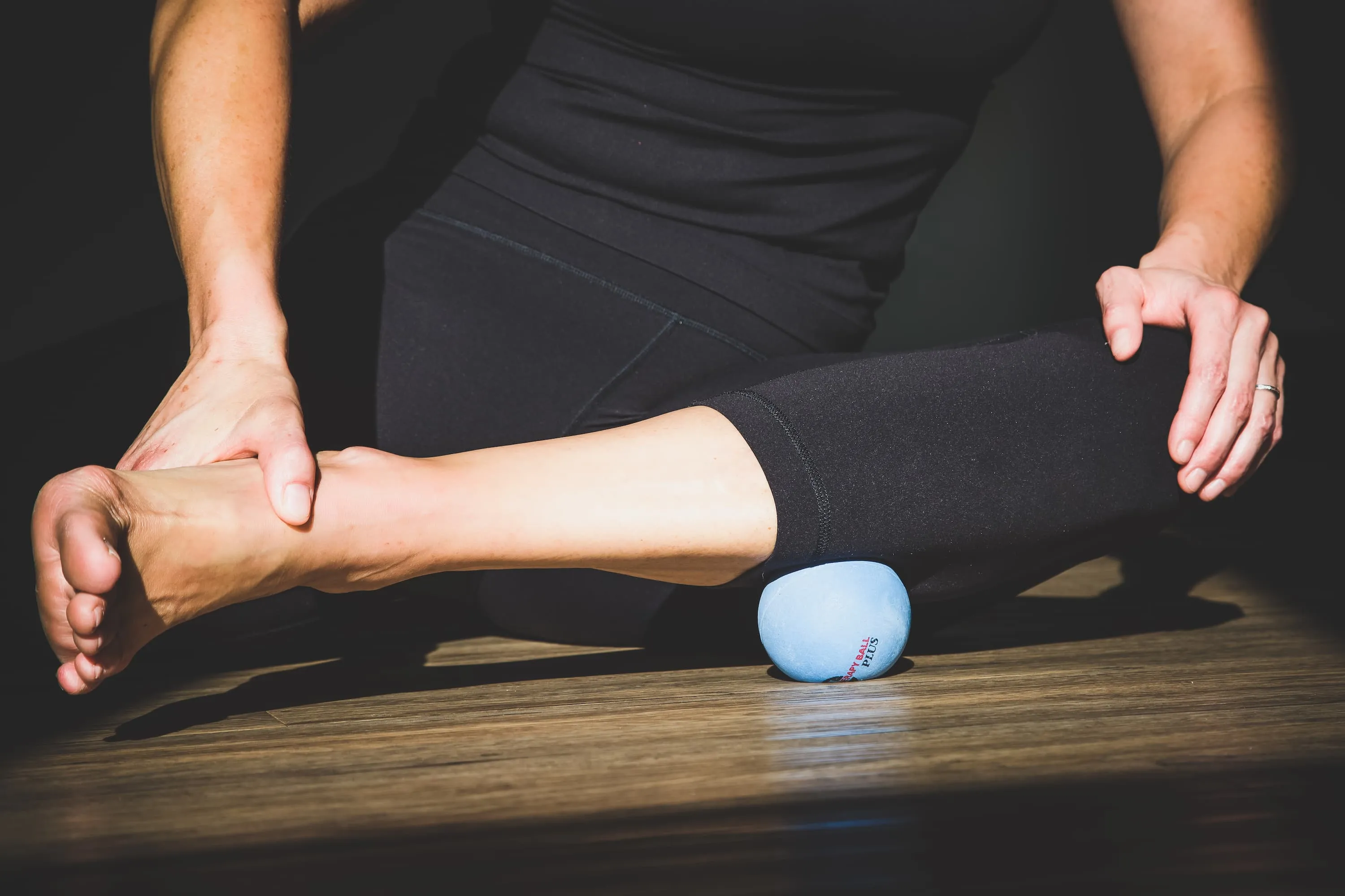 Lower body of woman with lower leg in front, hand holding foot, and therapy ball under calf muscle.