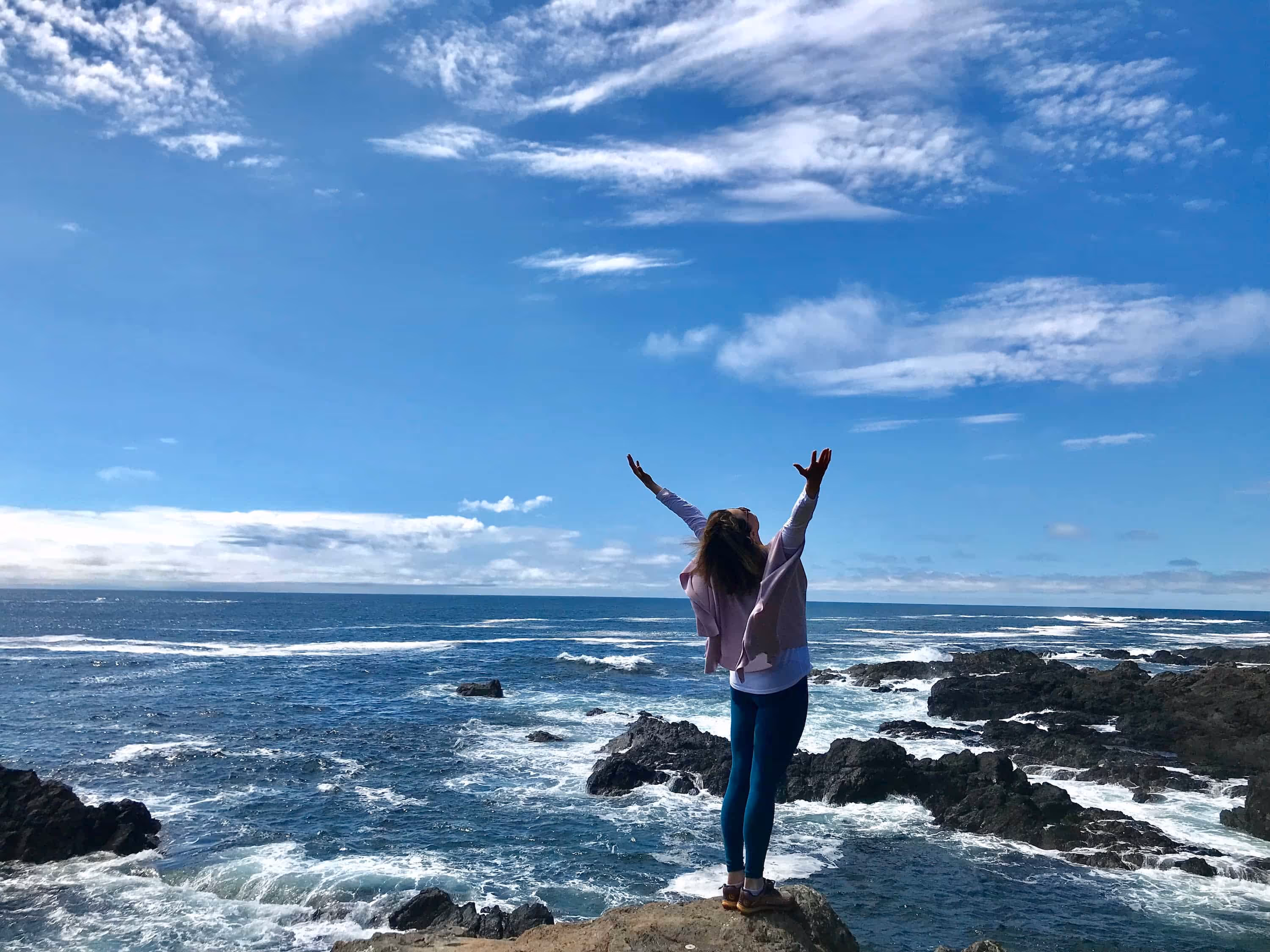 Women standing on rocks by the ocean with arms outstretched to sky.