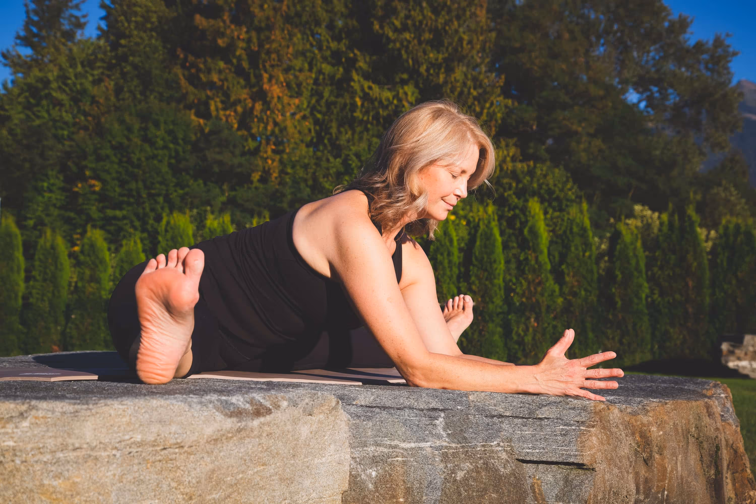 Sheri doing a seated wide leg forward fold on a large rock outside.