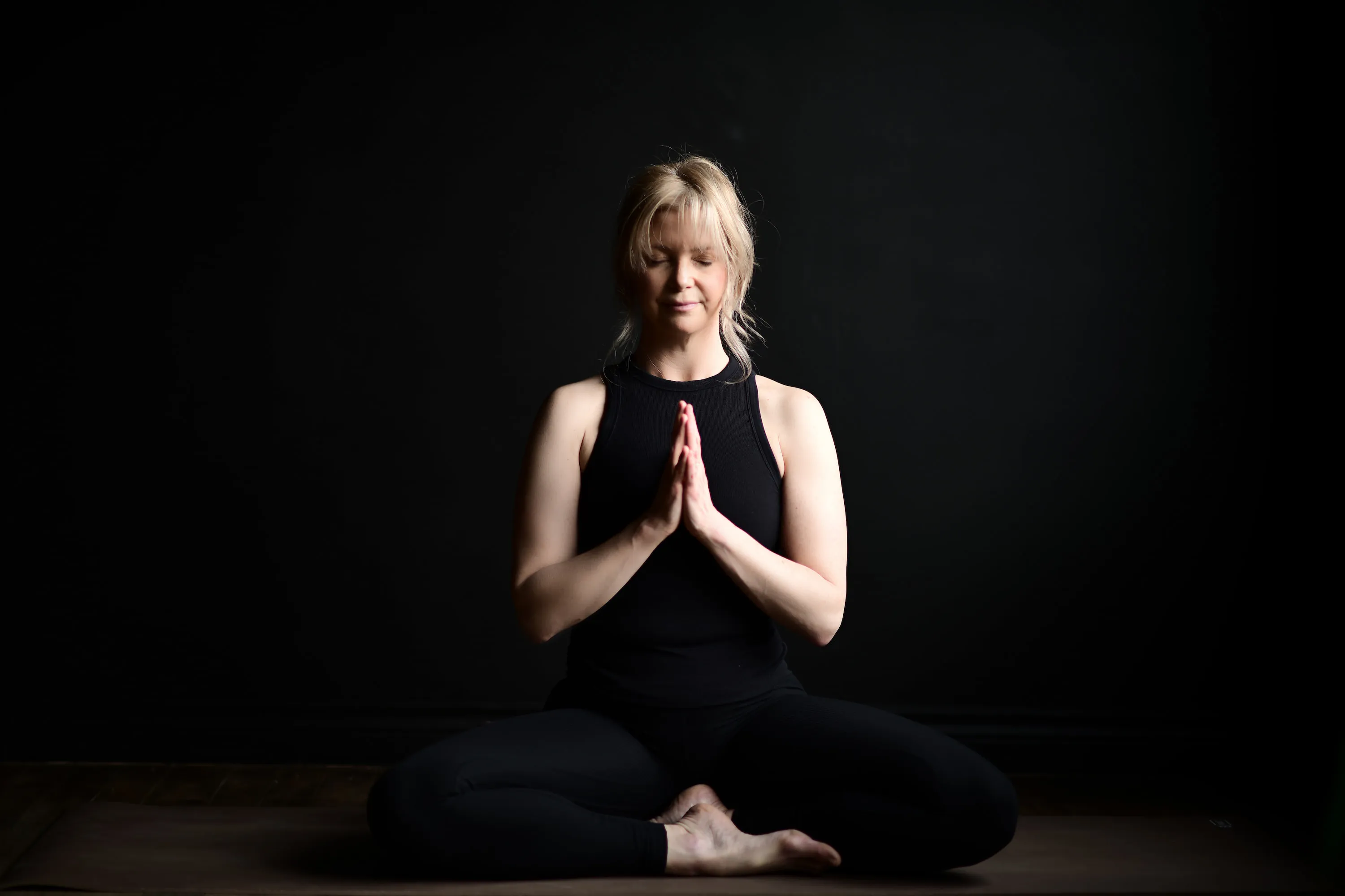 Sheri, dressed in black sitting in lotus pose, hands in prayer, eyes closed.