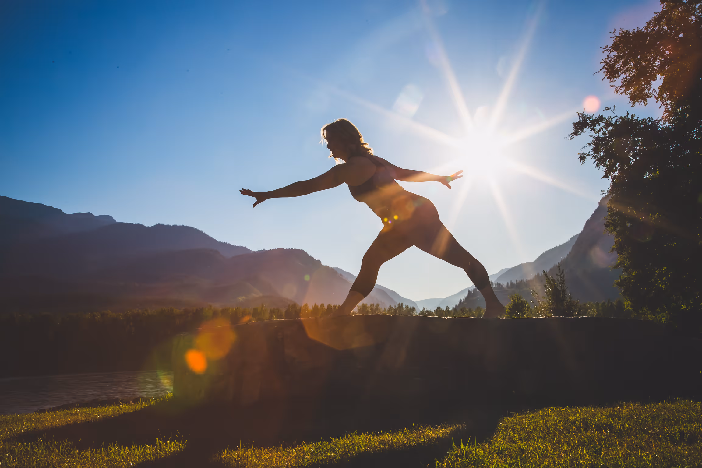 Lady standing outdoors on a rock, doing yoga pose. Sun and mountains shining behind her.