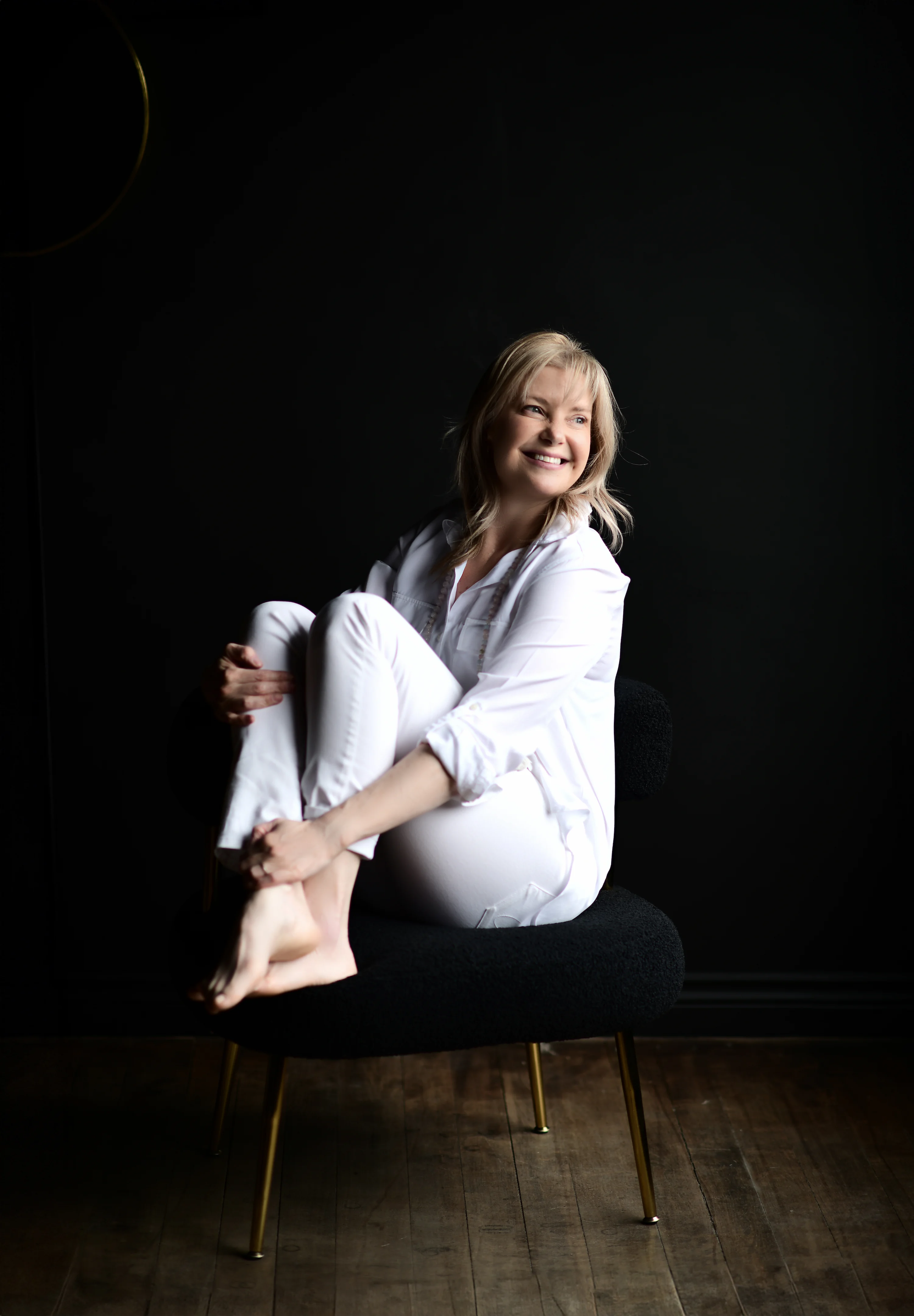 Sheri Zebroff dressed in white sitting on stool in dark room with wood floor.