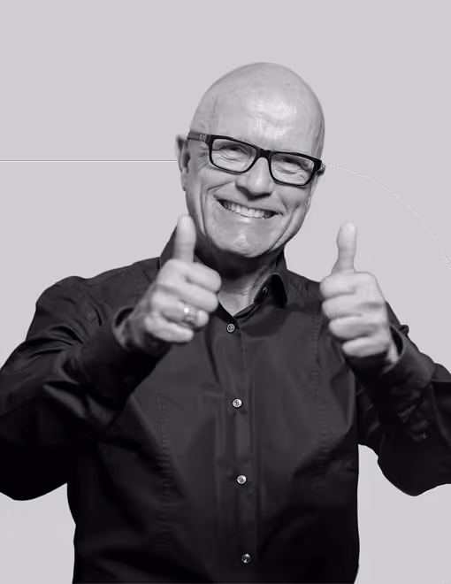 Mann mit Brille lächelt und zeigt Daumen hoch, trägt ein dunkles Hemd vor grauem Hintergrund.