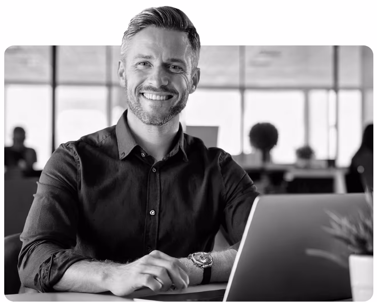 Mann mit Bart lächelt am Laptop, modernes Büro im Hintergrund.