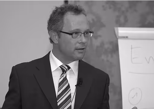 Mann im Anzug spricht vor Flipchart, trägt Brille und Krawatte, gestikuliert engagiert.
