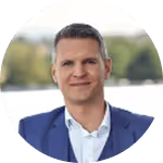 Ein Mann in einem blauen Anzug lächelt leicht. Der Hintergrund ist unscharf und zeigt grüne Bäume.