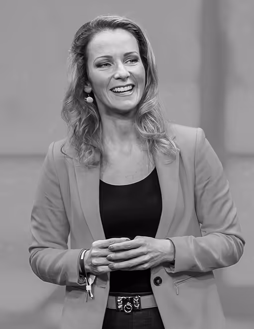 Frau lächelt in Blazer, redet auf Bühne, hält Hände zusammen.