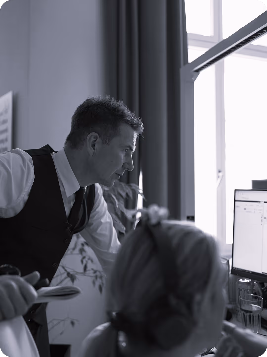 Mann in Weste arbeitet über Schulter einer Person an einem Computer, konzentriert, im Büro mit Pflanzen und großem Fenster.