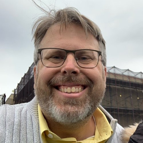 Christopher Tall smiling outdoors wearing glasses and sweater