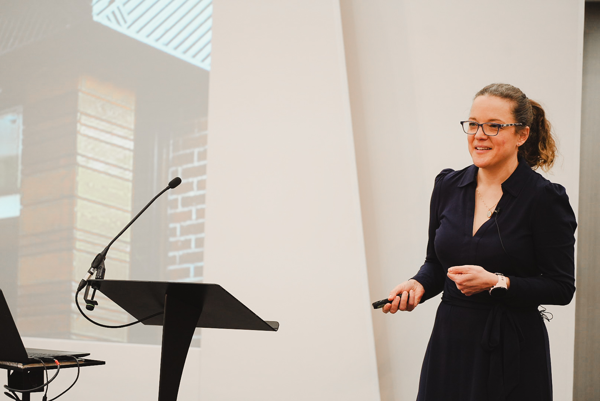 Woman in glasses speaking at a podium with a microphone and laptop during a presentation.