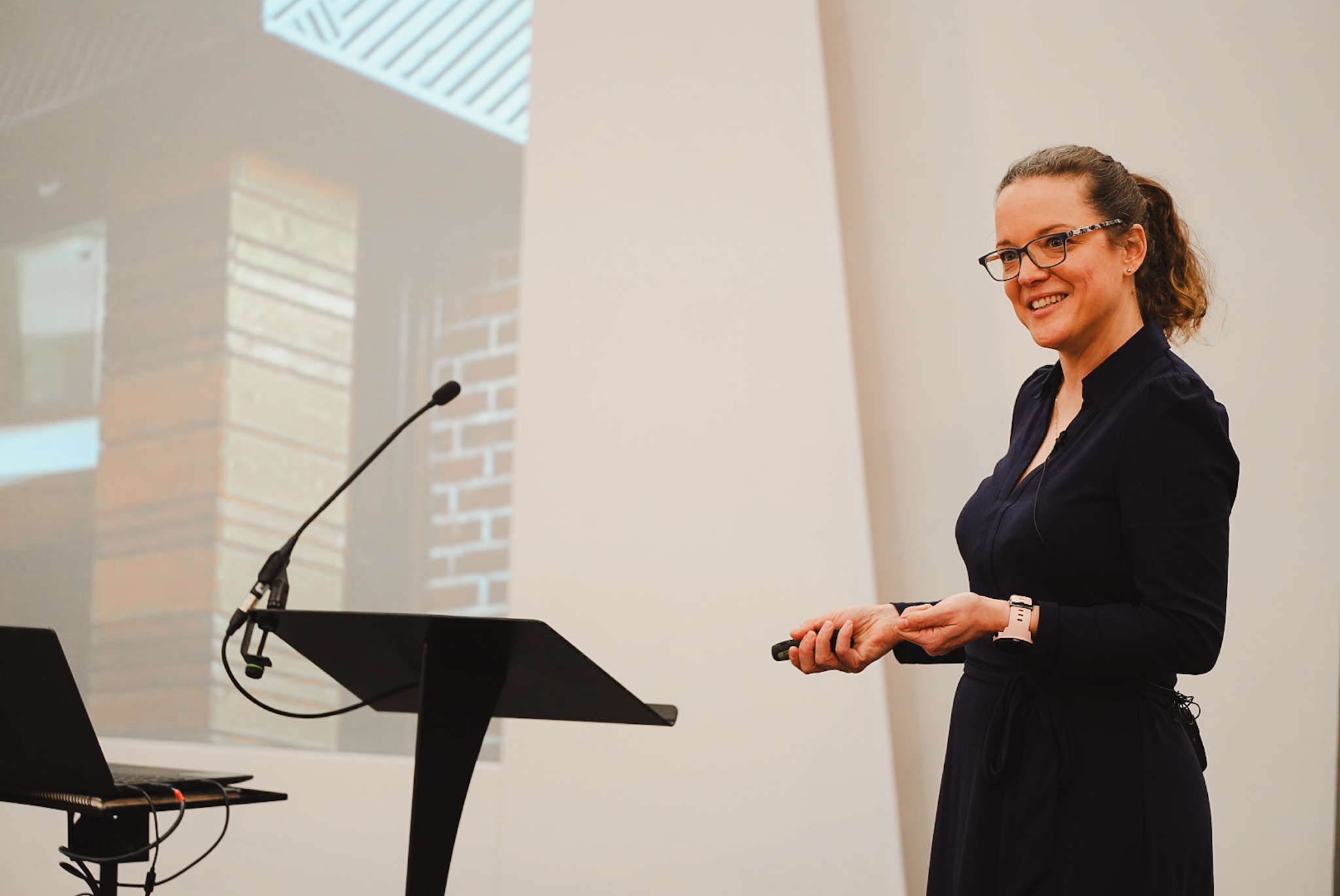 Woman with glasses smiles while giving a presentation next to a microphone and laptop on a podium.