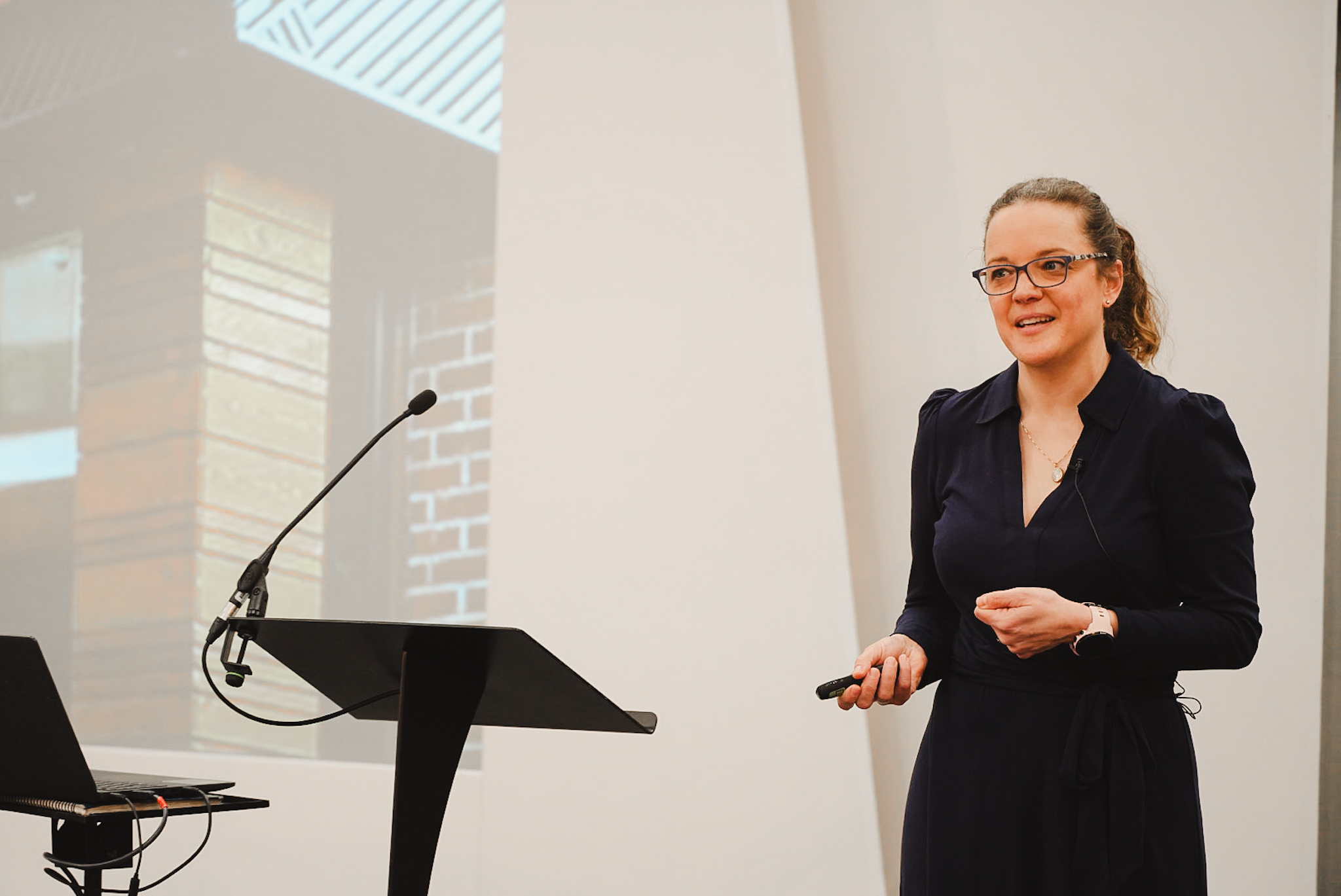 Woman in glasses and dark dress speaking at a podium with a microphone and laptop nearby.