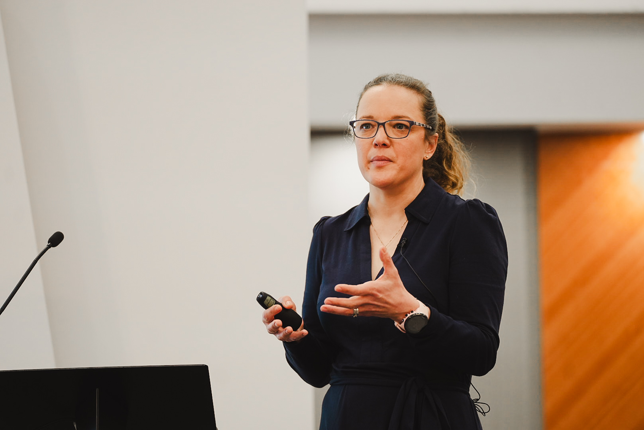 Woman with glasses and ponytail giving a presentation using a clicker and standing near a microphone.