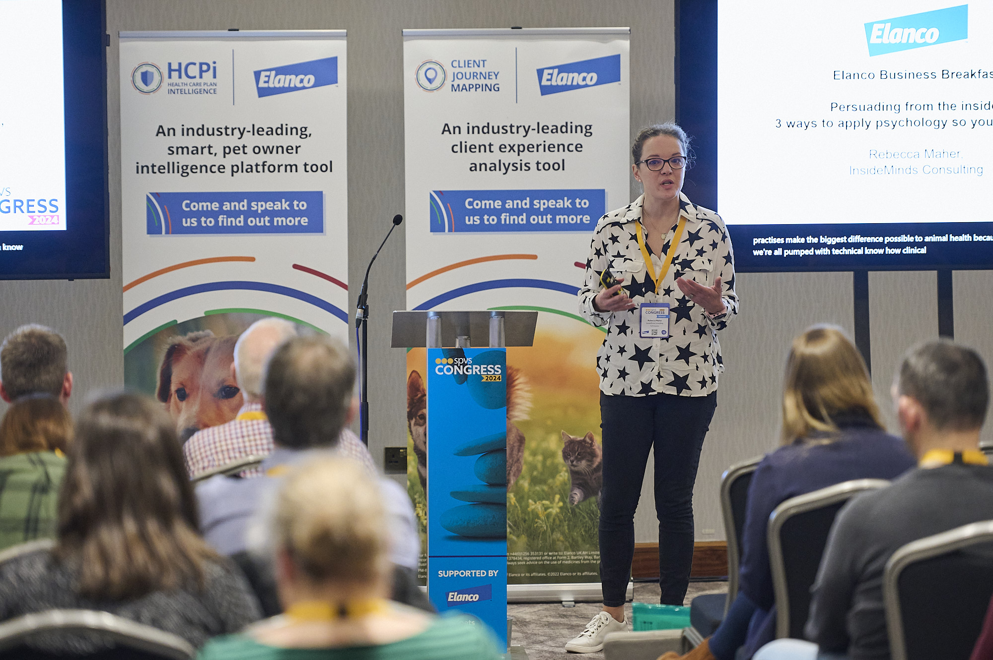 Woman speaker with star-patterned shirt presents at a conference with banners and audience visible.