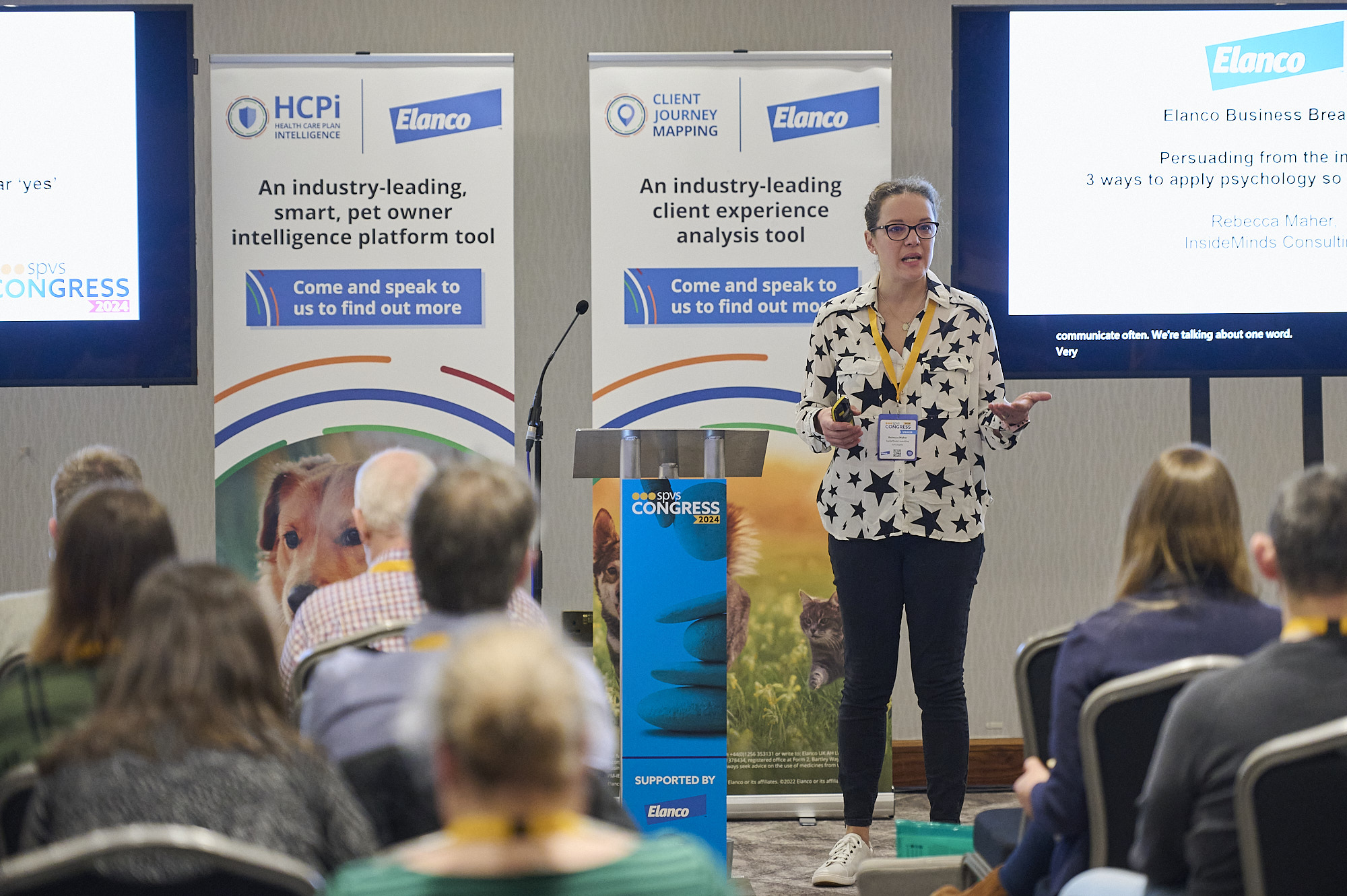 Woman in star-patterned shirt speaks to seated audience at a pet care conference presentation.