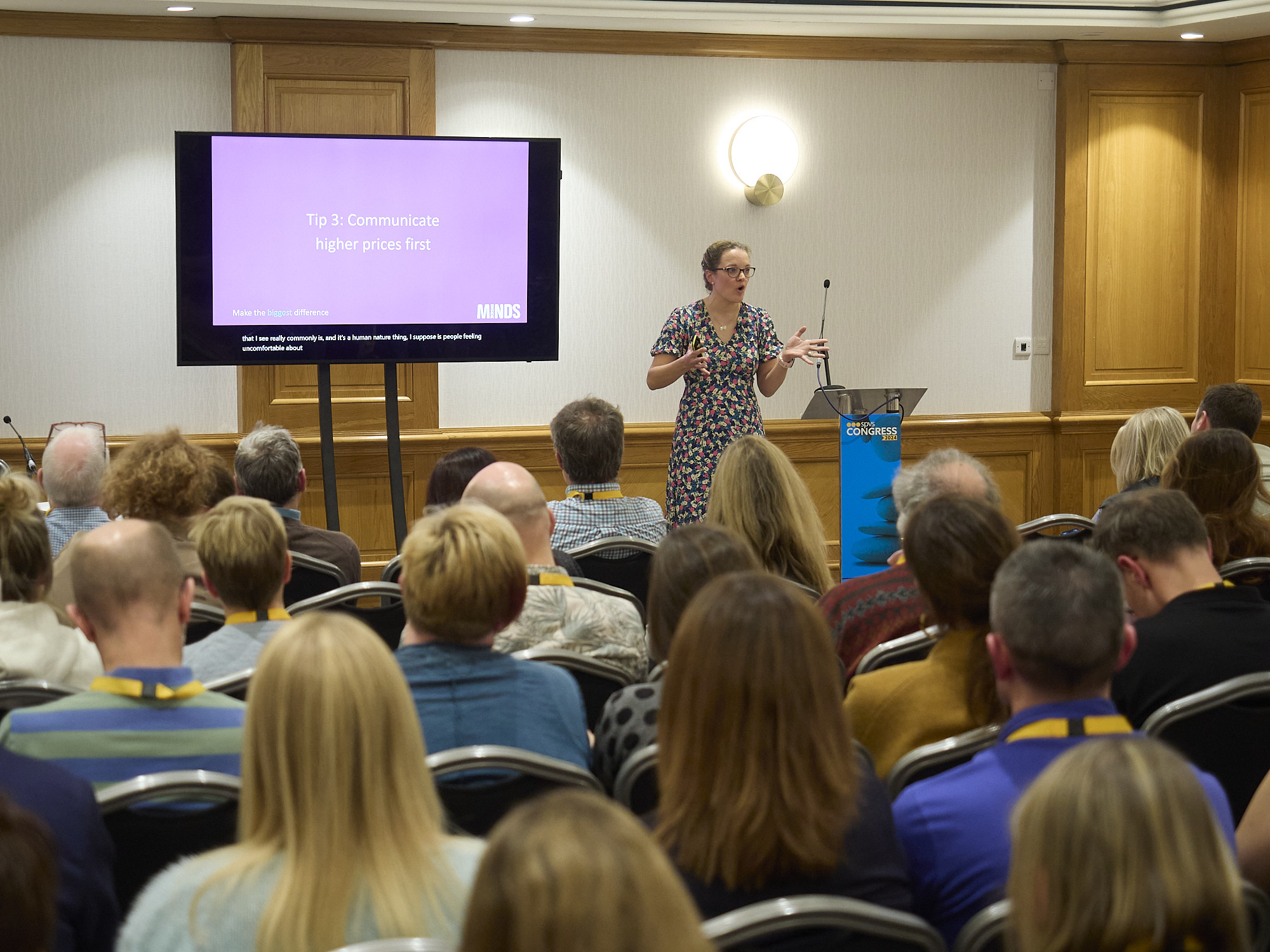 Woman presenting Tip 3: Communicate higher prices first to seated audience at a conference.