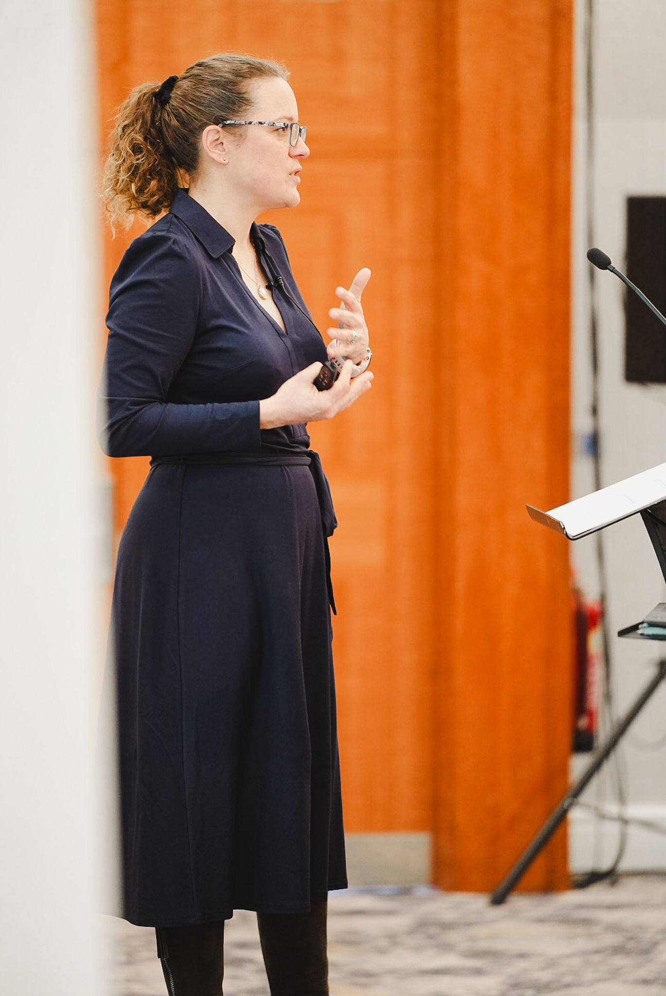 Woman with glasses and curly hair speaks in front of a microphone with an orange wall background.