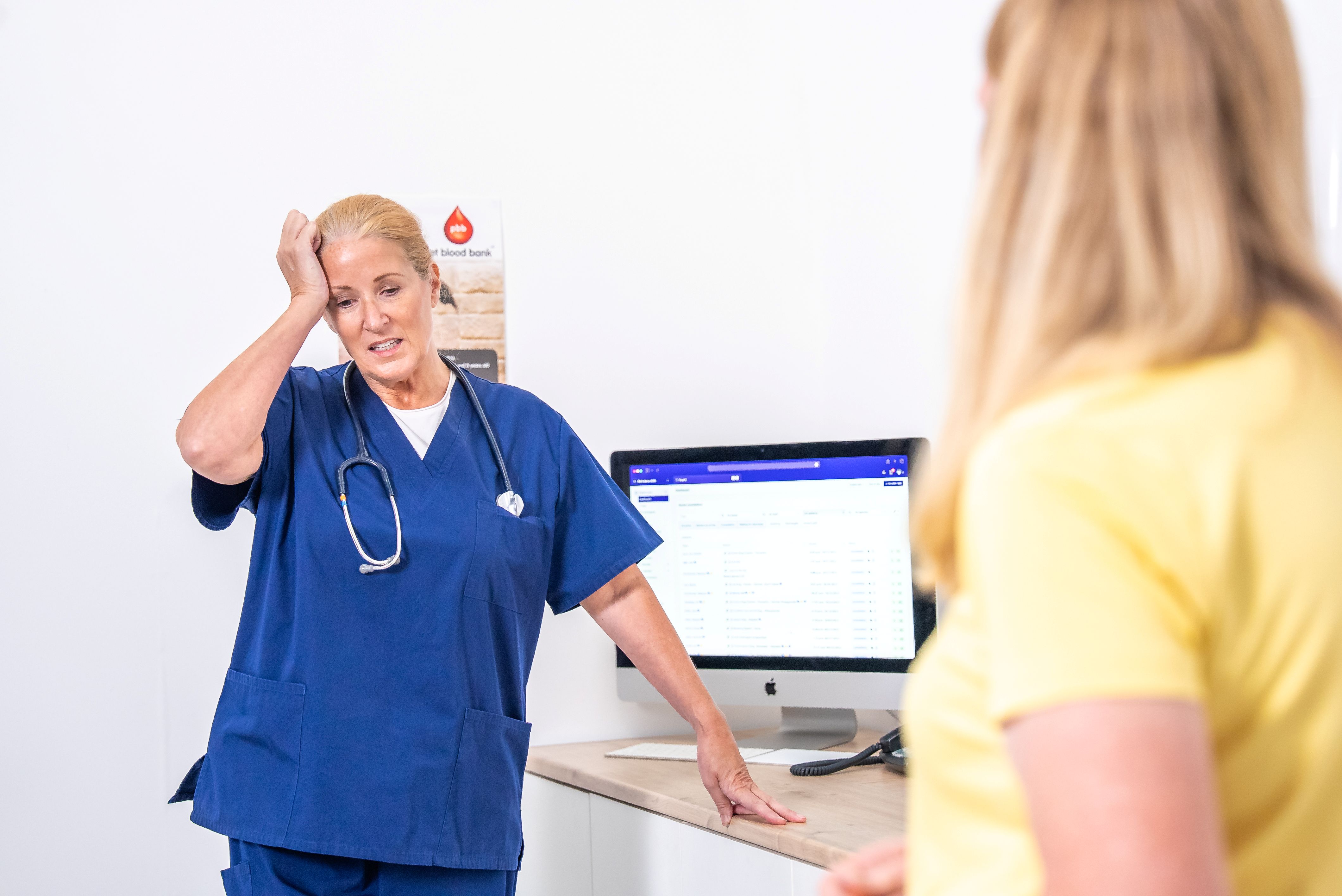 Tired nurse in blue scrubs leaning on desk, speaking to a person in a yellow shirt in a clinic room.