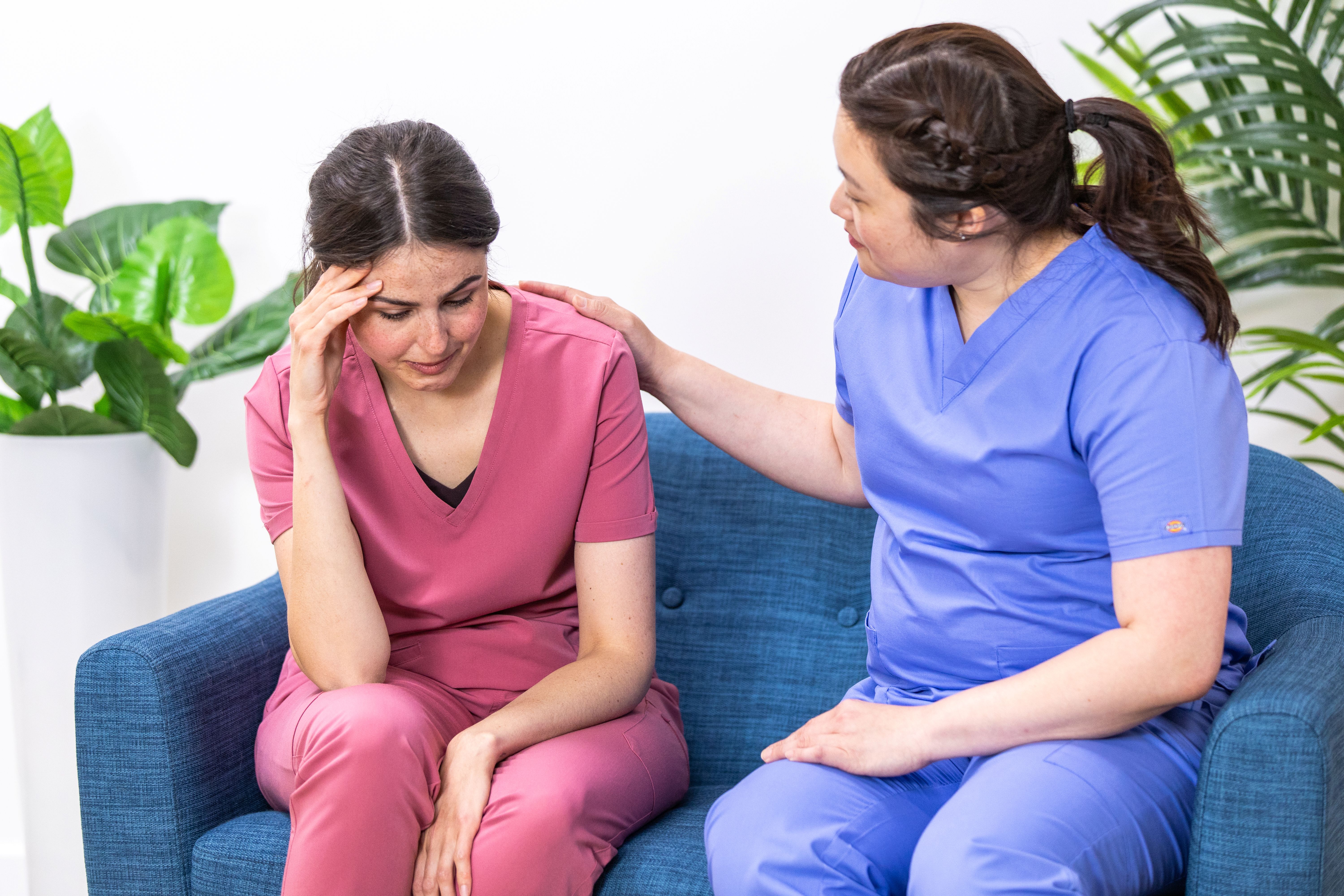 Healthcare worker in blue scrubs comforting a distressed colleague in pink scrubs on a blue couch.
