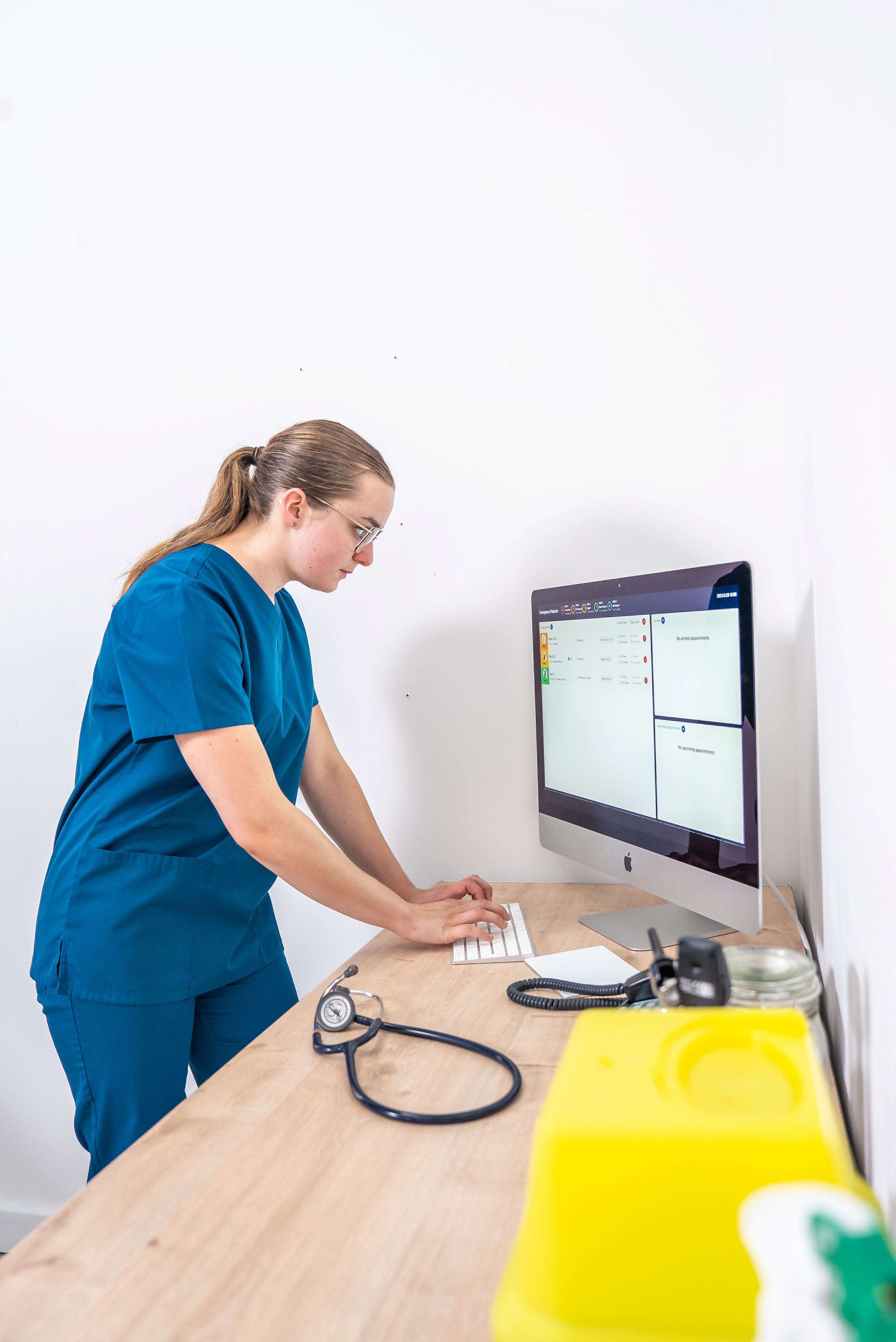 Medical professional in blue scrubs typing on a computer at a wooden desk with stethoscope nearby.
