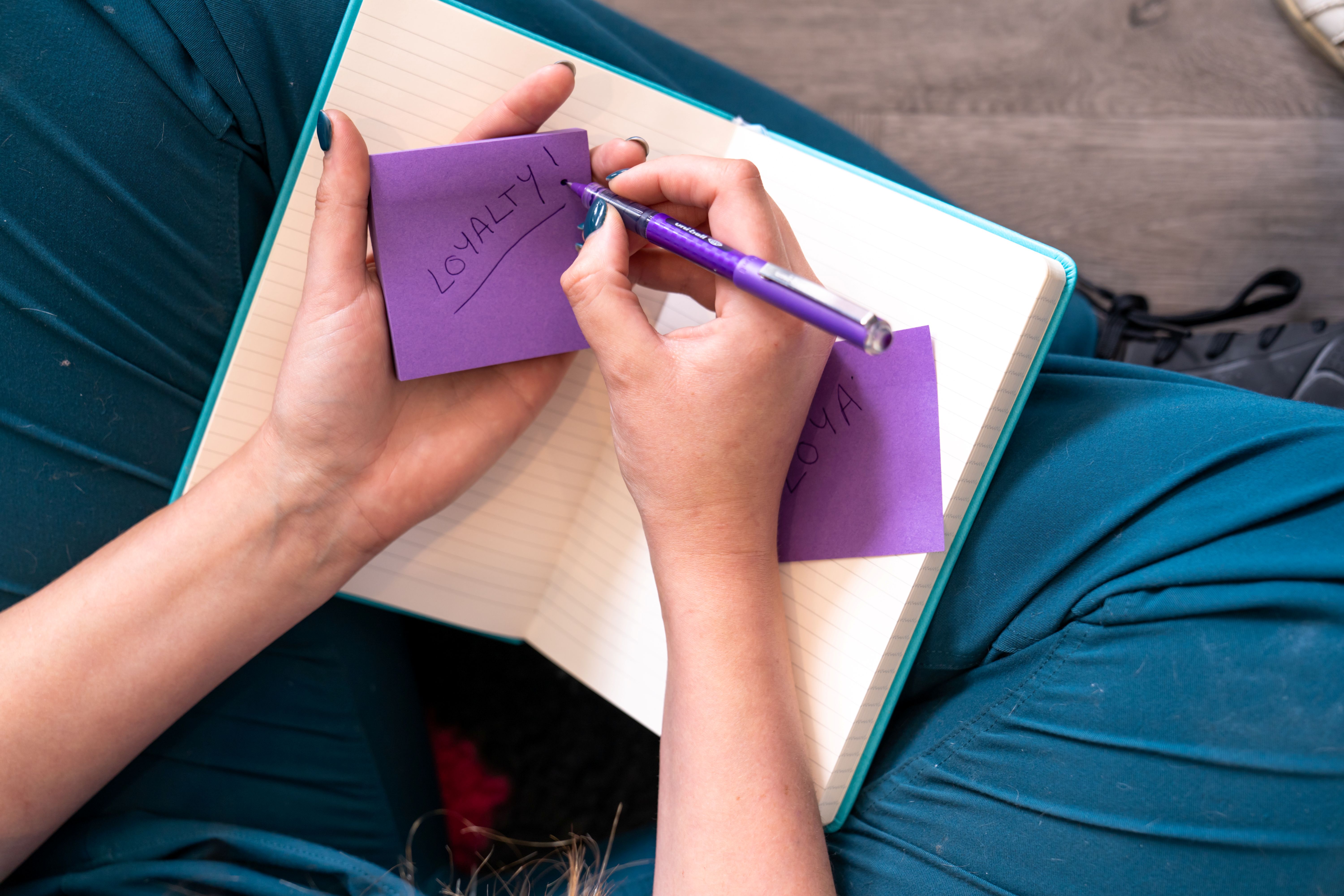 Person writing the word 'LOYALTY!' on a purple sticky note with a purple pen over a notebook.