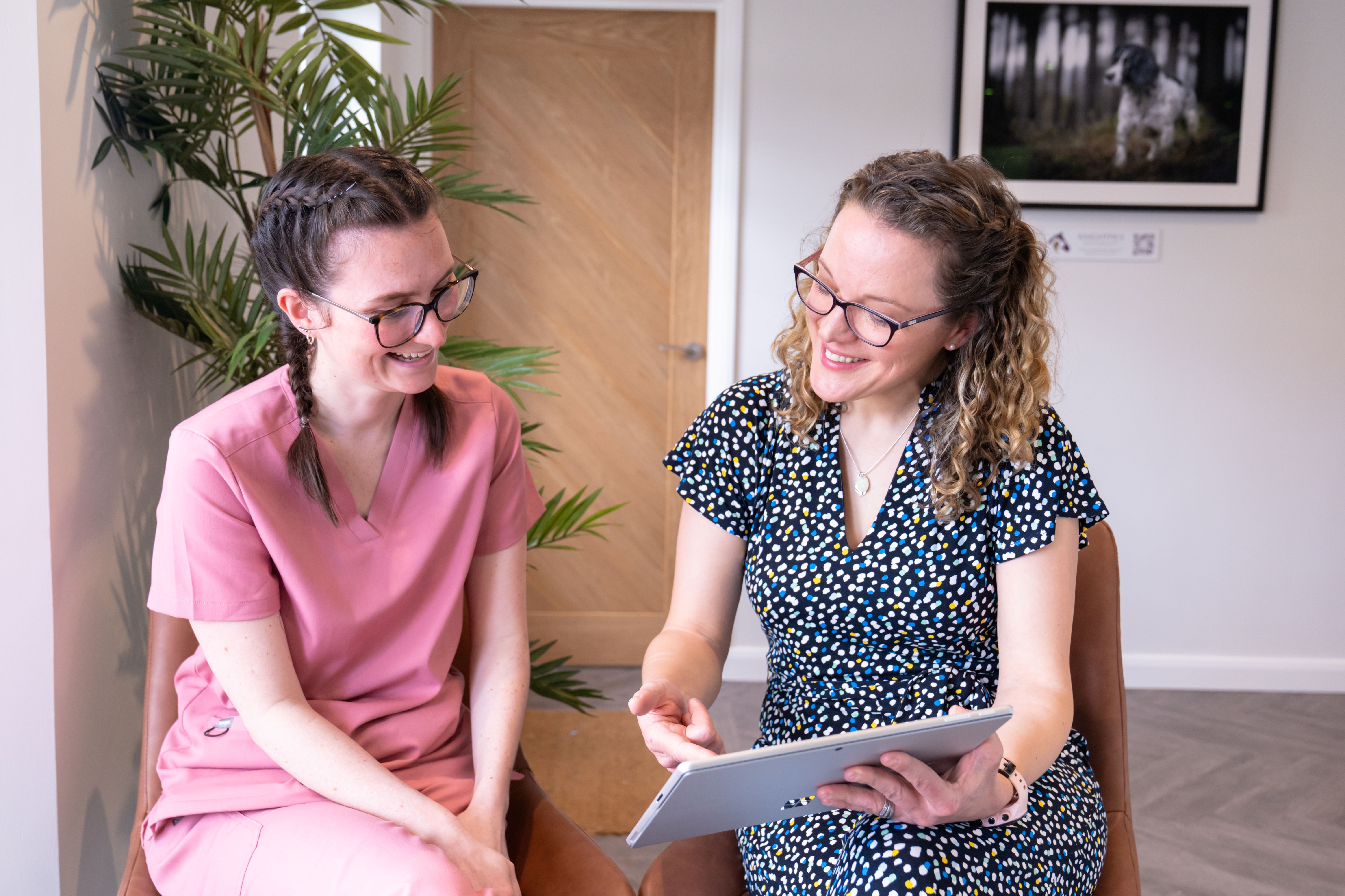 Two women with glasses smiling and looking at a tablet while seated in a bright room.