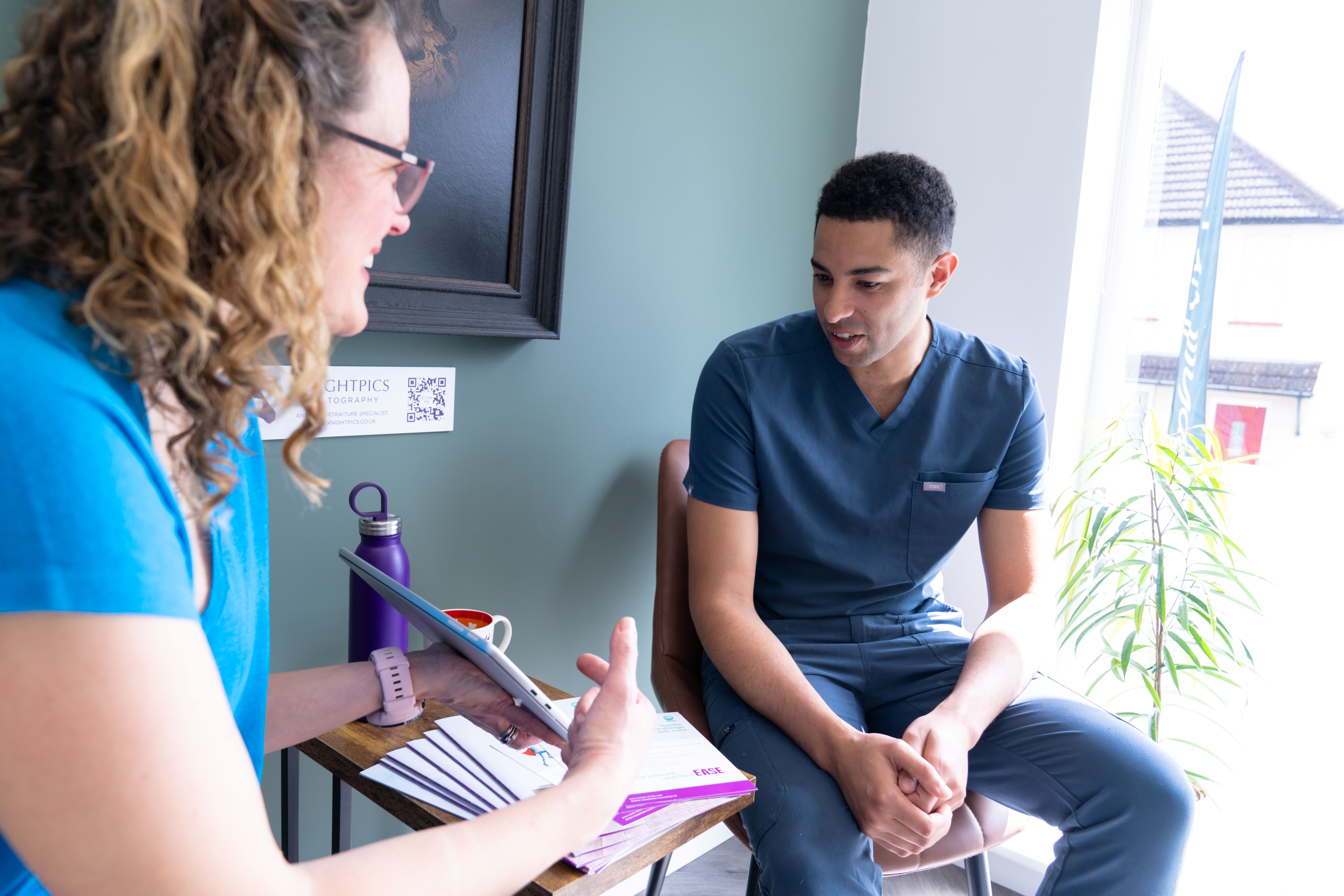 A woman in blue scrubs shows a tablet to a man in blue scrubs sitting by a window with a plant.