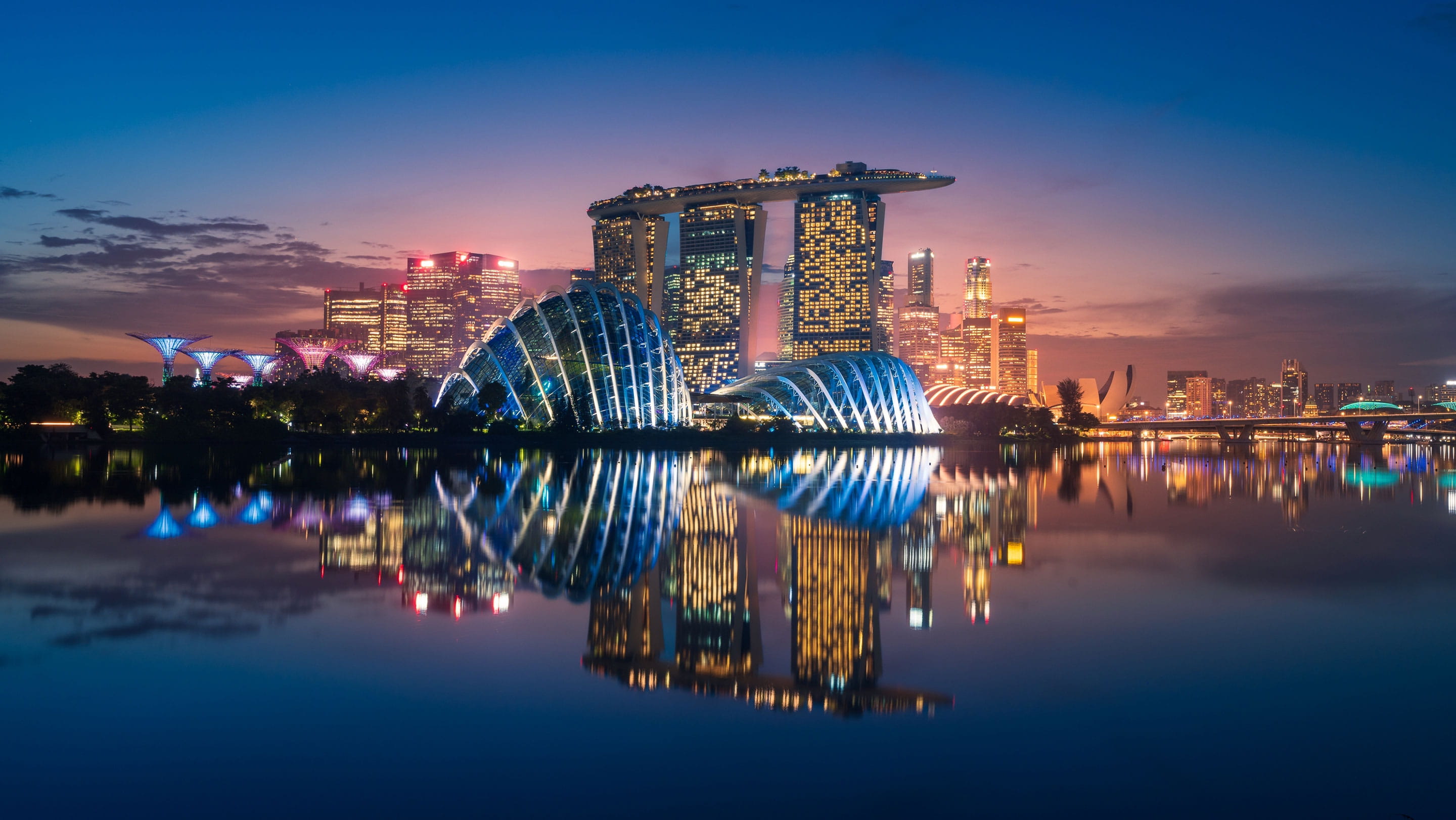 Singapore's Marina Bay skyline at sunset, featuring Marina Bay Sands, Gardens by the Bay and illuminated skyscrapers reflected in the water.