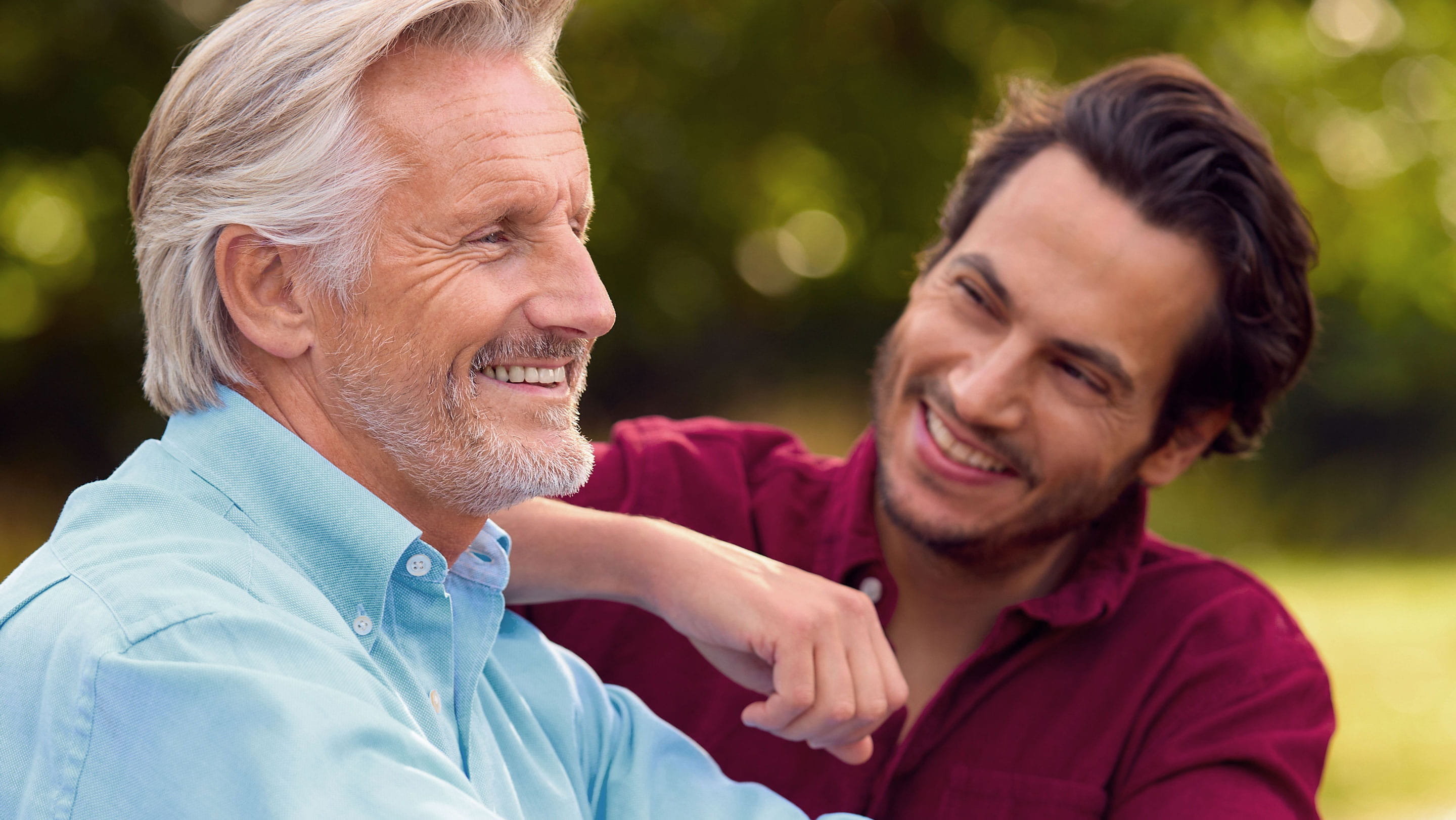 An older man with grey hair smiling alongside a younger man in an outdoor setting, illustrating intergenerational relationships, family succession and the transfer of wealth.