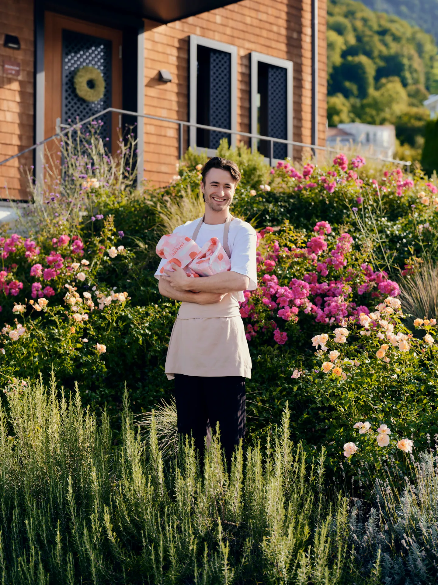 Ein lächelnder Mann mit beigem Schürzenhemd steht in einem Garten voller blühender Rosen in Rosa- und Aprikosenfarben vor einem Haus mit Holzverkleidung und hält mehrere rosa-weiße Päckchen mit der Aufschrift „Bäckerei“ in den Armen.