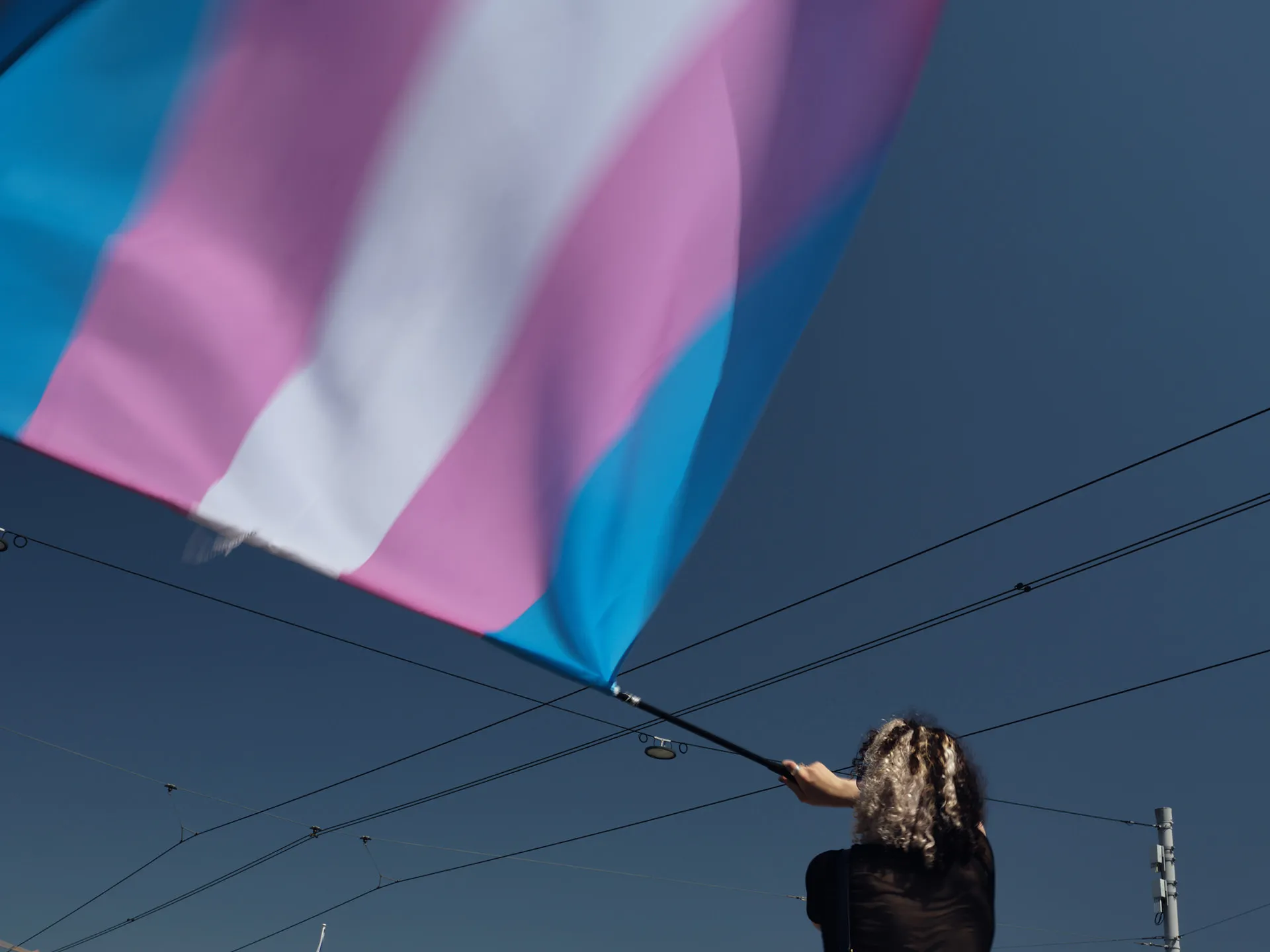 Person waving a large transgender pride flag with blue, pink, and white stripes against a clear blue sky with overhead wires.