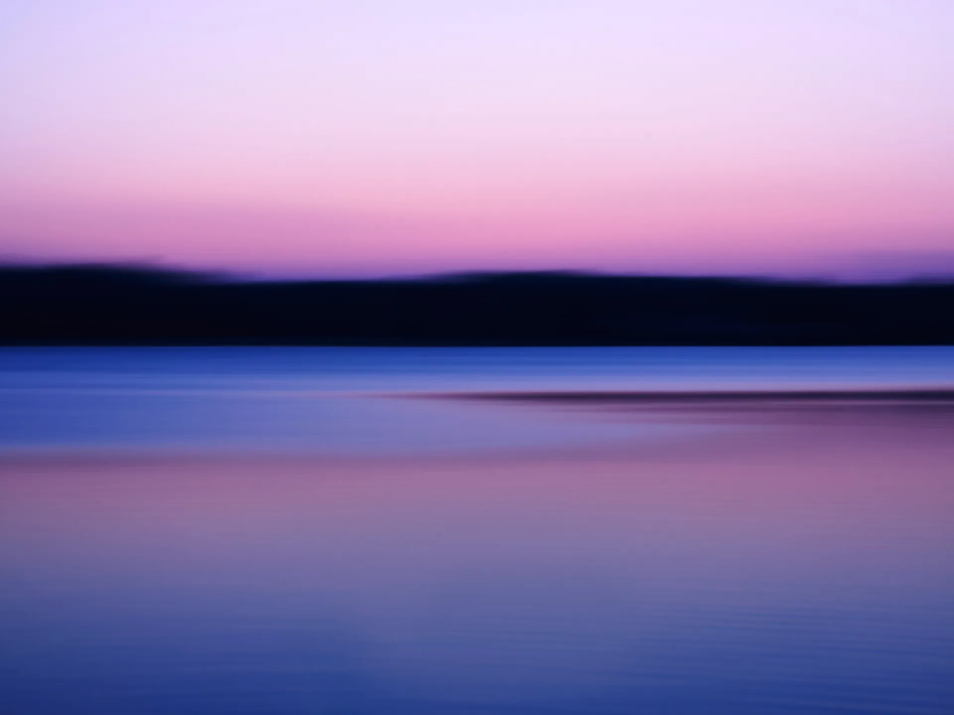 Verschwommene Landschaft mit dunklem Horizont, blauem Wasser und Himmel im Rosa-Lila-Farbverlauf bei Dämmerung.