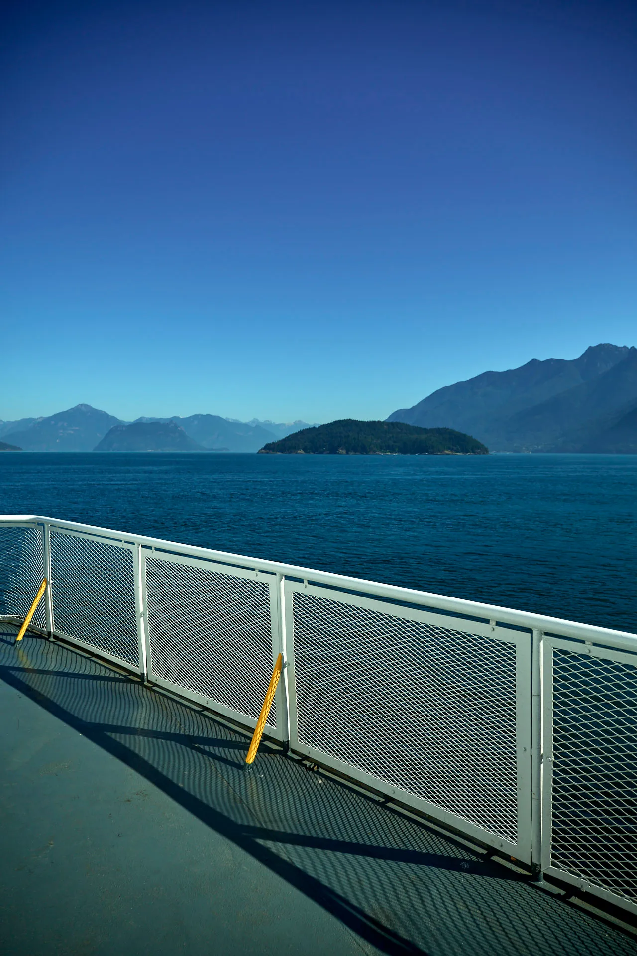 Blick von der Reling einer Fähre auf einen ruhigen blauen See mit einer bewaldeten Insel und Bergen im Hintergrund unter klarem Himmel.