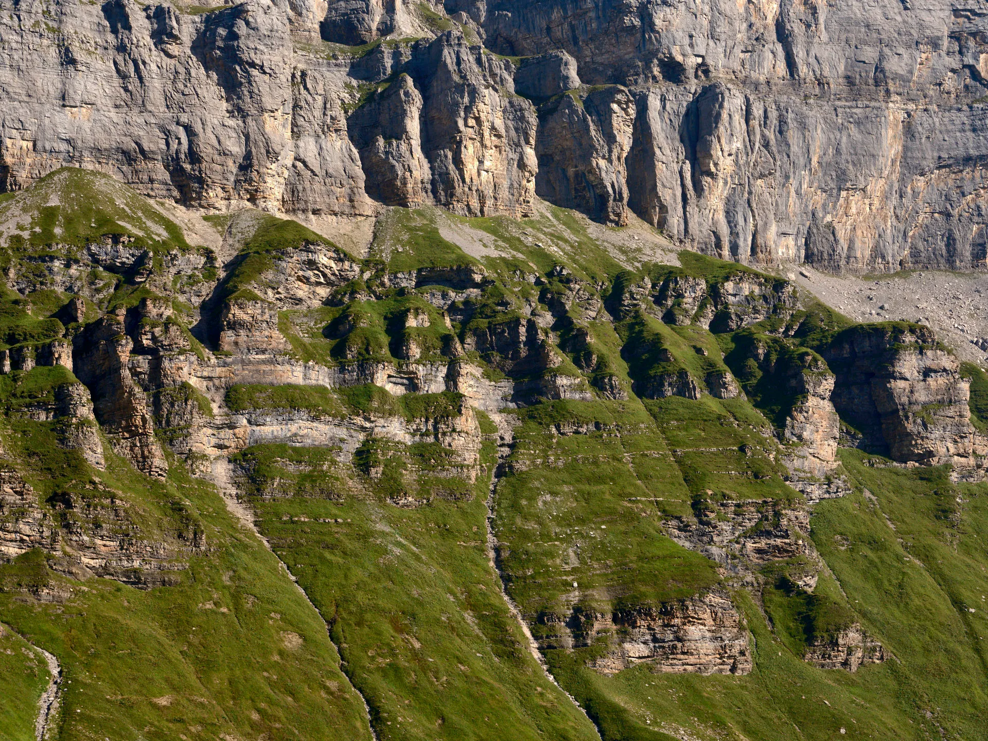 Steile Felswand mit zahlreichen Gesteinsschichten und bewachsenen Grasflächen in sonnigem Gelände.