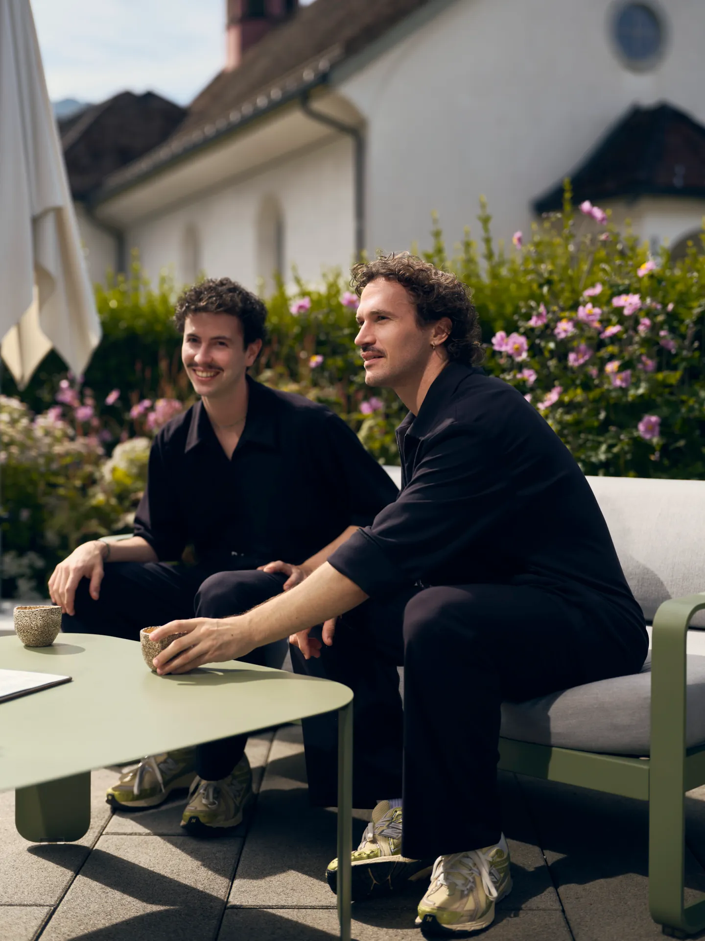 Zwei Männer mit lockigem Haar und dunkler Kleidung sitzen draußen auf einer Terrasse auf einem grauen Sofa neben einem grünen Tisch mit zwei Tassen, von Blumen und einer weißen Hauswand umgeben.