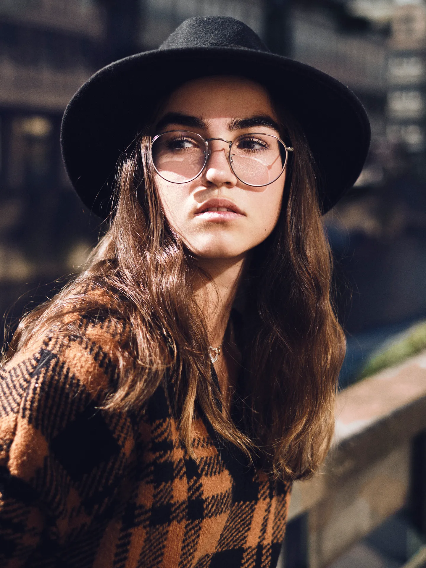 Junge Frau mit langen braunen Haaren, die einen schwarzen Hut, runde Brille und einen braun-schwarz karierten Pullover trägt, blickt nach rechts. Das Bild zeigt sie im Freien mit unscharfem Hintergrund.