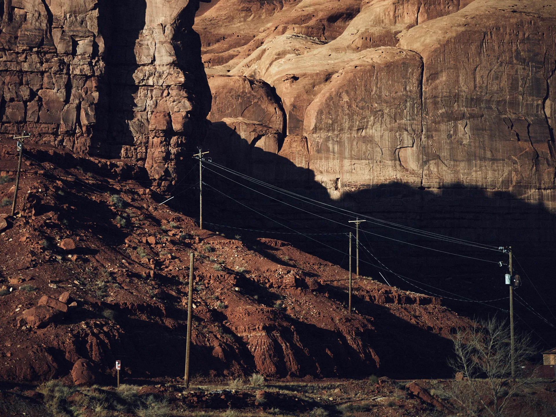 Rote Felsenformationen bei Sonnenlicht mit langen Schatten und Strommasten auf felsigem Boden vor den Klippen.