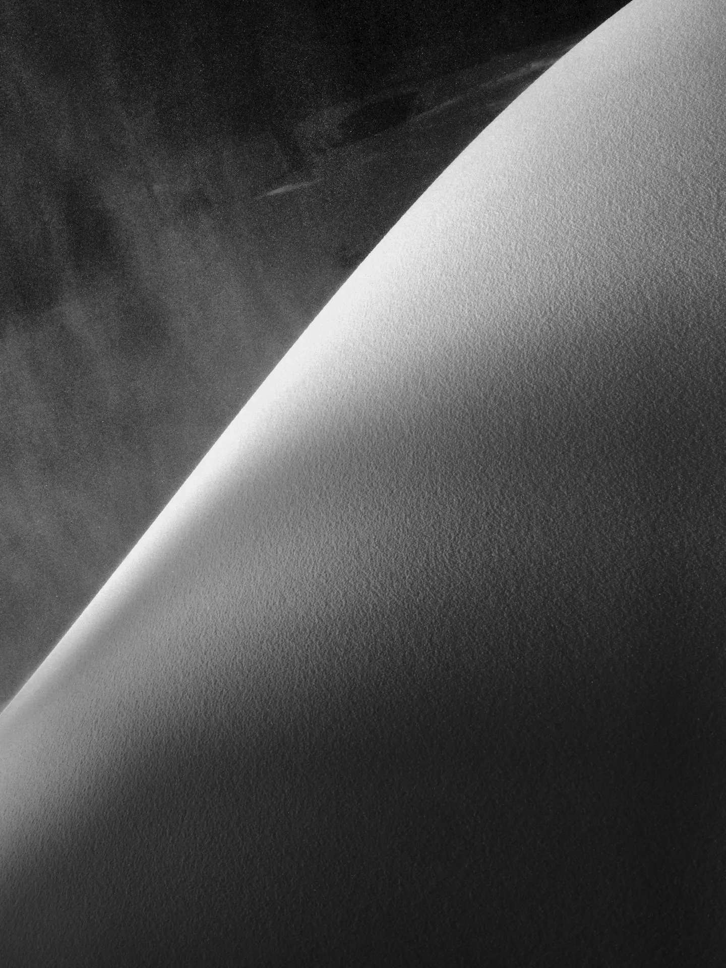 Schwarz-weiss-Bild einer schräg verlaufenden, glatten Planetenoberfläche, die von hellem Licht von links oben beleuchtet wird, mit dunklem, nebelartigen Weltraum im Hintergrund.