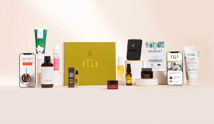 Coffret Box by ELLE x Claris Virot présenté sur fond beige, comprenant une sélection de produits de beauté, soins du corps et du visage, compléments alimentaires, accessoires et contenus numériques. Au centre, une boîte dorée estampillée « Box by ELLE ». Deux smartphones affichent des contenus numériques liés à la box. L'ensemble est soigneusement disposé sur une surface claire.