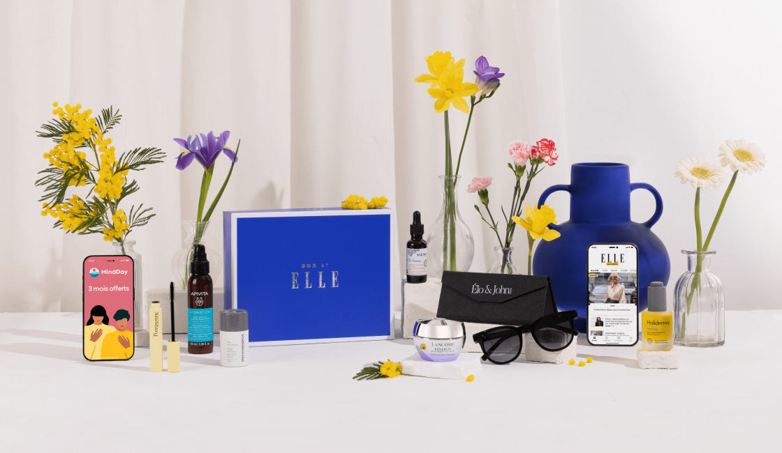 Coffret Box by ELLE x Elo & John présenté sur une table blanche avec un fond crème, entouré de fleurs fraîches en vase. Au centre, une boîte bleue siglée Box by ELLE. Autour, divers produits cosmétiques et bien-être : soins du visage, sérum, crème Lancôme, vernis à ongles, mascara, ainsi qu’une paire de lunettes de soleil dans un étui Elo & John. Deux smartphones affichent les applis MindDay et ELLE.fr.