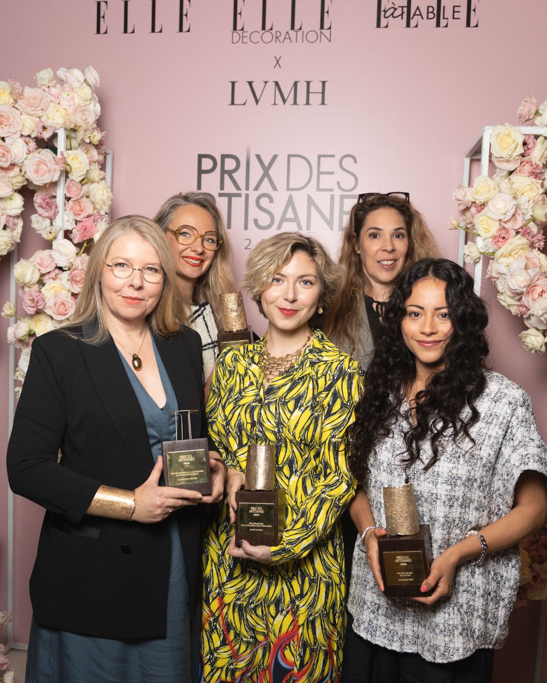 Cinq femmes posent devant un mur fleuri rose pastel décoré des logos ELLE, ELLE Décoration, ELLE à Table et LVMH, sous l’inscription « Prix des Artisanes 2024 ». Trois d’entre elles tiennent un trophée en bois et métal doré. Toutes sourient, dans une ambiance conviviale. La photo célèbre les lauréates de cette édition du prix récompensant des femmes engagées dans les métiers de l’artisanat.