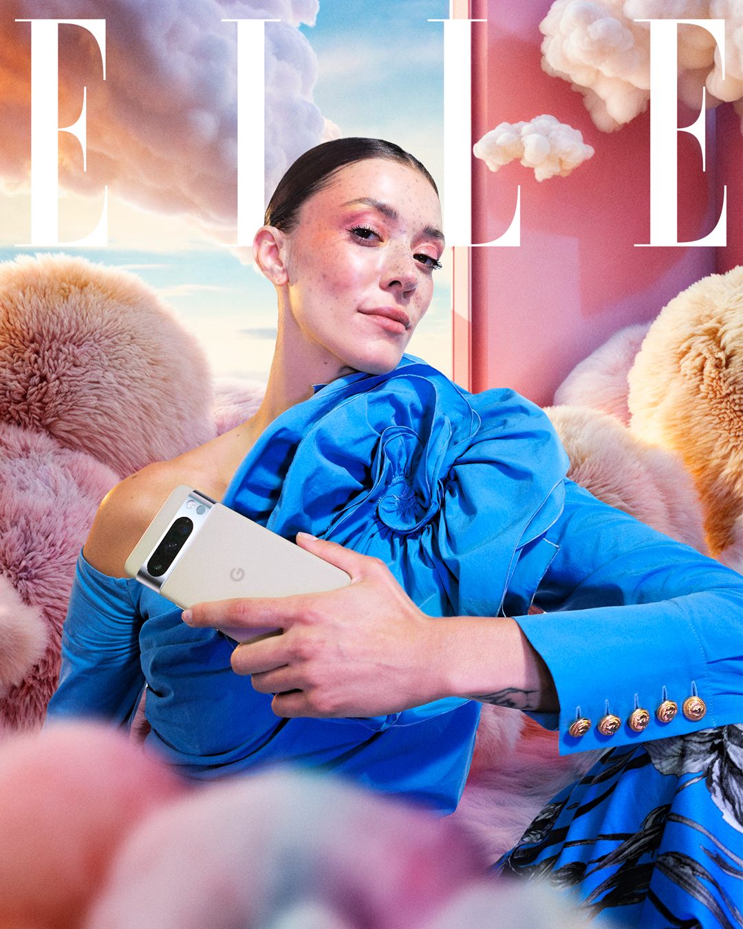 Femme assise dans un décor onirique aux tons pastel, entourée de coussins en fausse fourrure rose et beige et de nuages suspendus. Elle porte une tenue haute couture bleu vif avec des détails volumineux sur l'épaule et des boutons dorés sur la manche. Elle tient dans sa main un smartphone Google Pixel. Le logo "ELLE" apparaît en grand en haut de l'image, dans un ciel de coucher de soleil stylisé.