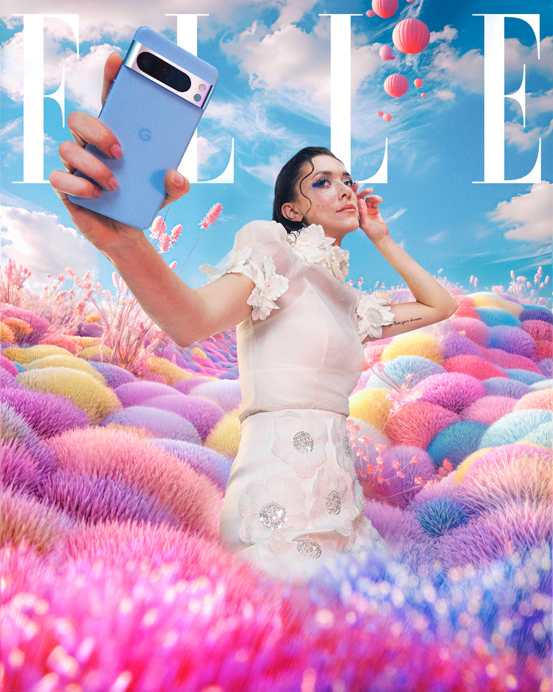 Femme debout dans un champ imaginaire de touffes végétales aux couleurs pastel (rose, violet, jaune, bleu). Elle porte une robe blanche ornée de fleurs en relief, lève un smartphone Google Pixel bleu dans sa main gauche pour prendre un selfie, et regarde vers l’horizon. Son maquillage artistique inclut des paillettes autour des yeux. Des montgolfières flottent dans un ciel bleu vif. Le logo "ELLE" apparaît en blanc en haut de l’image.
