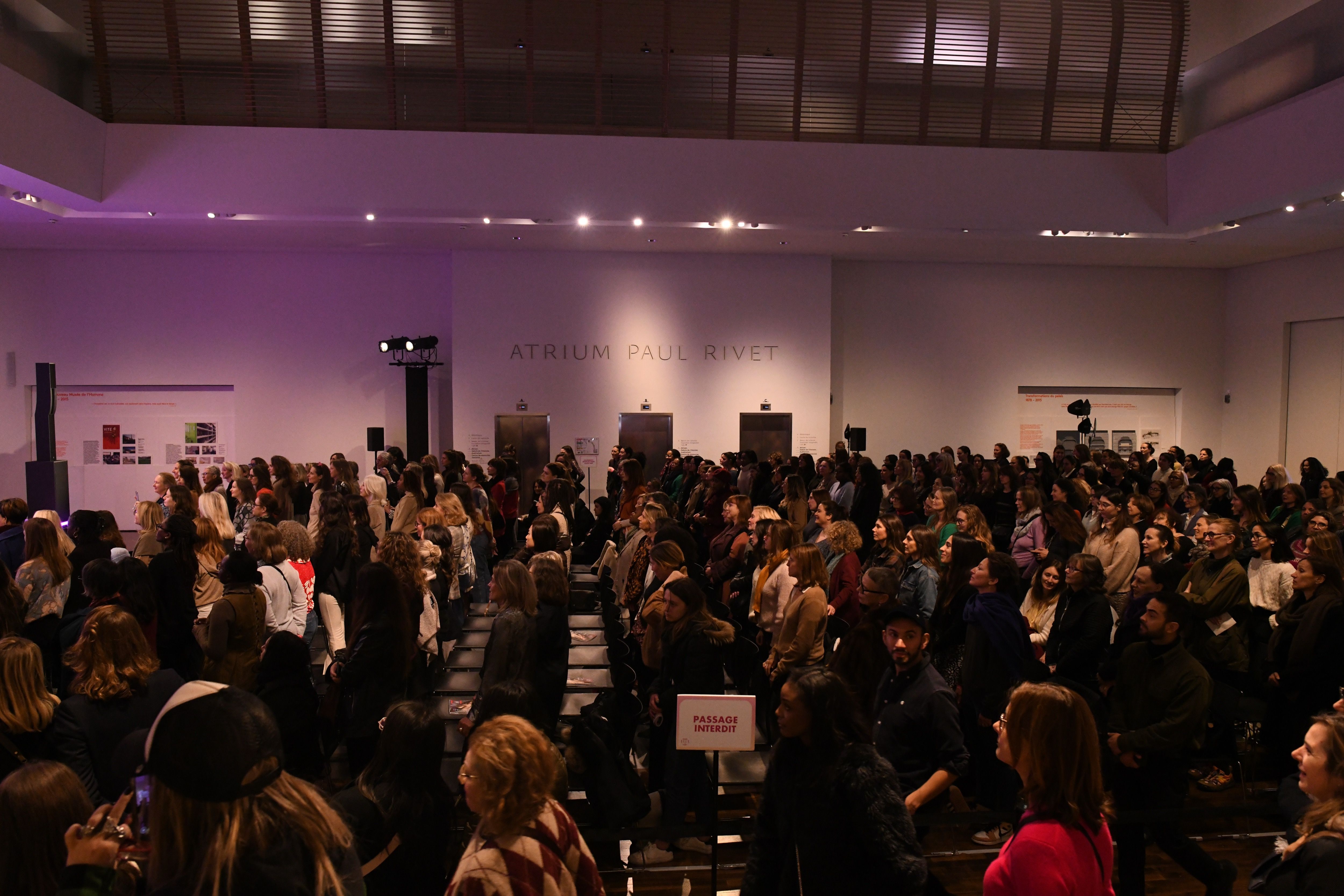 Vue d'ensemble d'une grande salle de conférence, l’Atrium Paul Rivet, remplie de personnes debout, attentives à un événement. Le public, majoritairement composé de femmes, regarde vers une scène hors champ. La salle est spacieuse, éclairée par des lumières tamisées, et des panneaux d’information sont visibles sur les murs. Un panneau « Passage interdit » est placé au centre au premier plan.